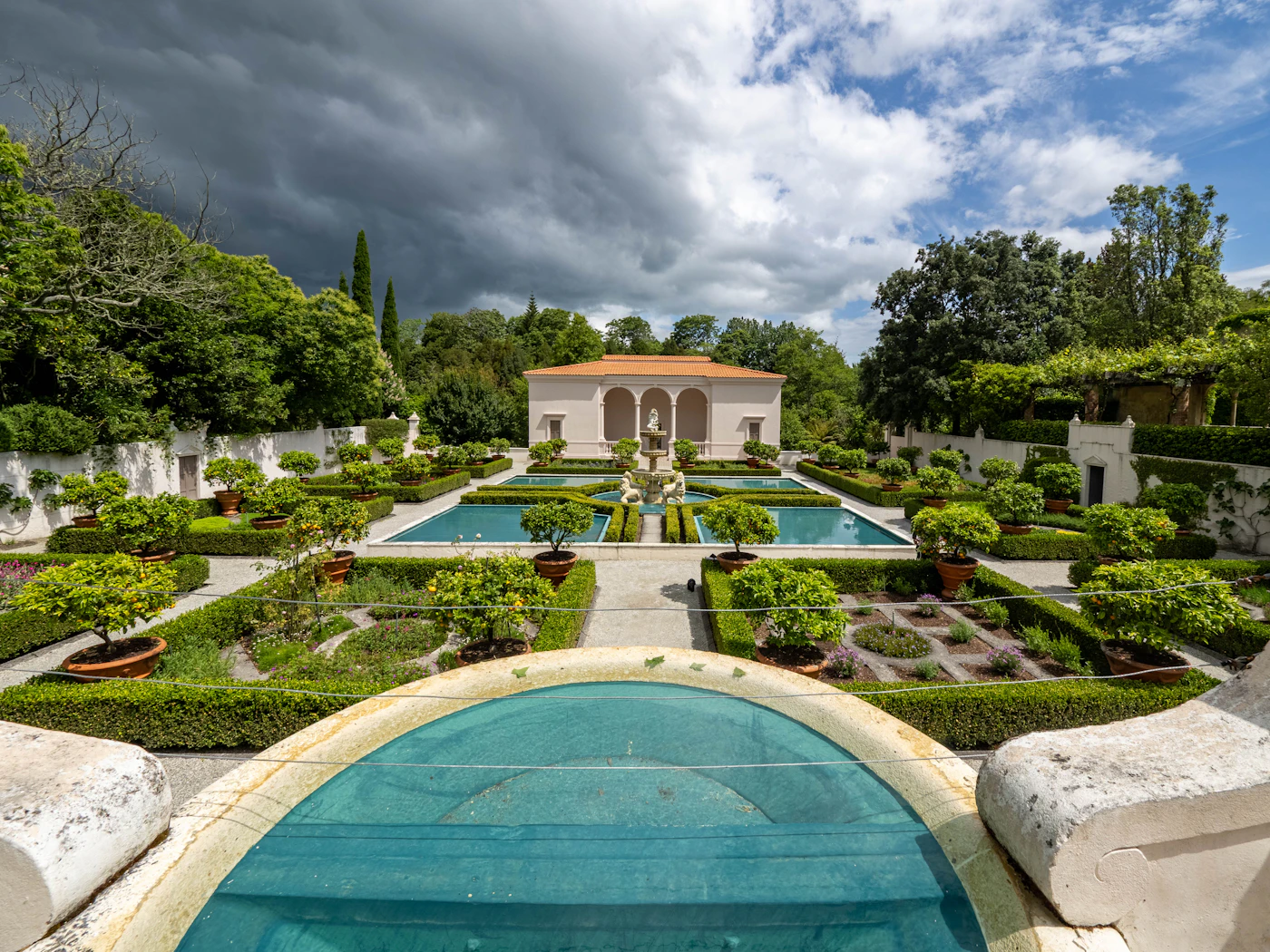 A formal garden with clipped hedges, gravel paths and a reflecting pool in front of a stone residence.