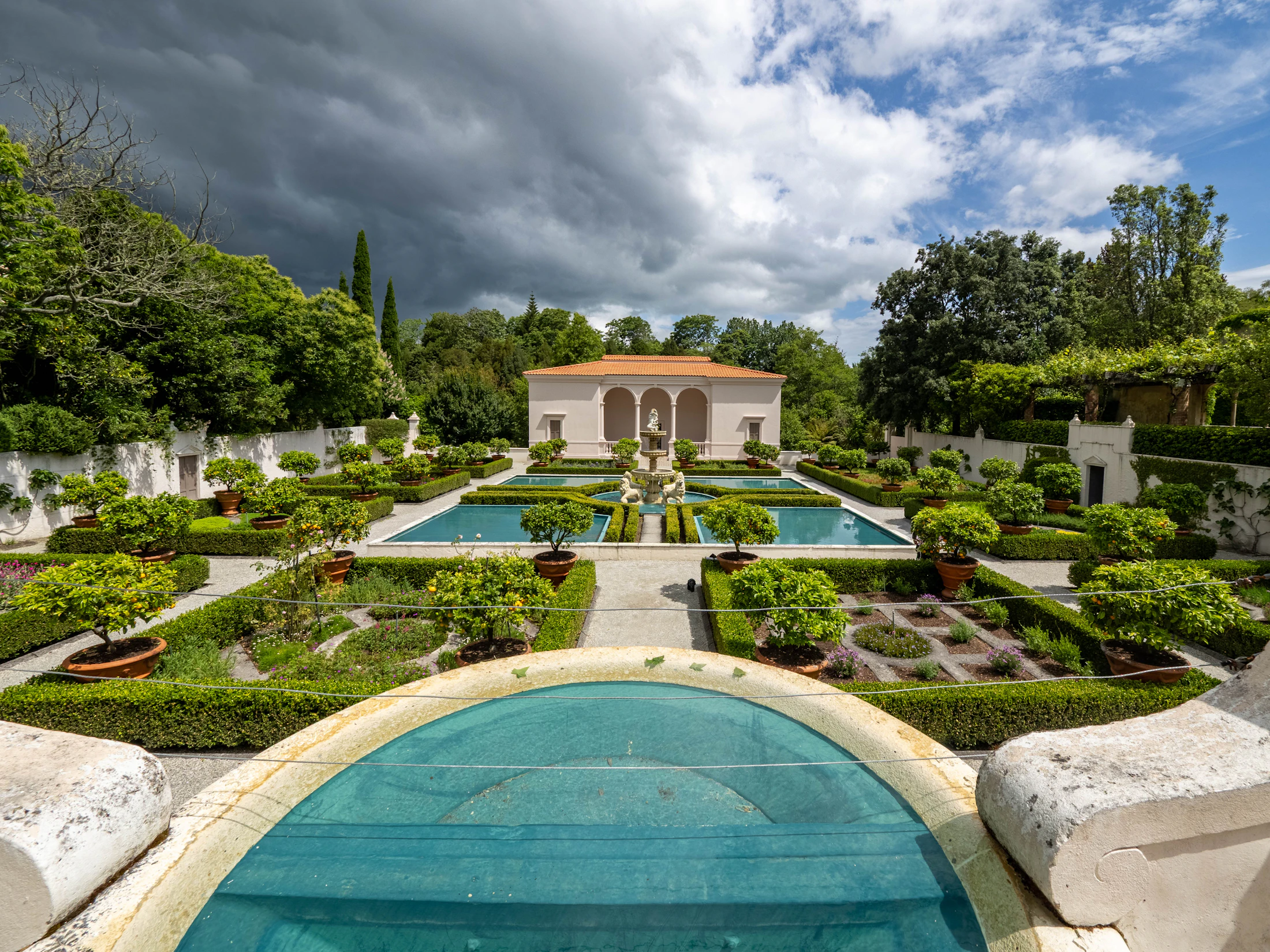 A formal garden with clipped hedges, gravel paths and a reflecting pool in front of a stone residence.