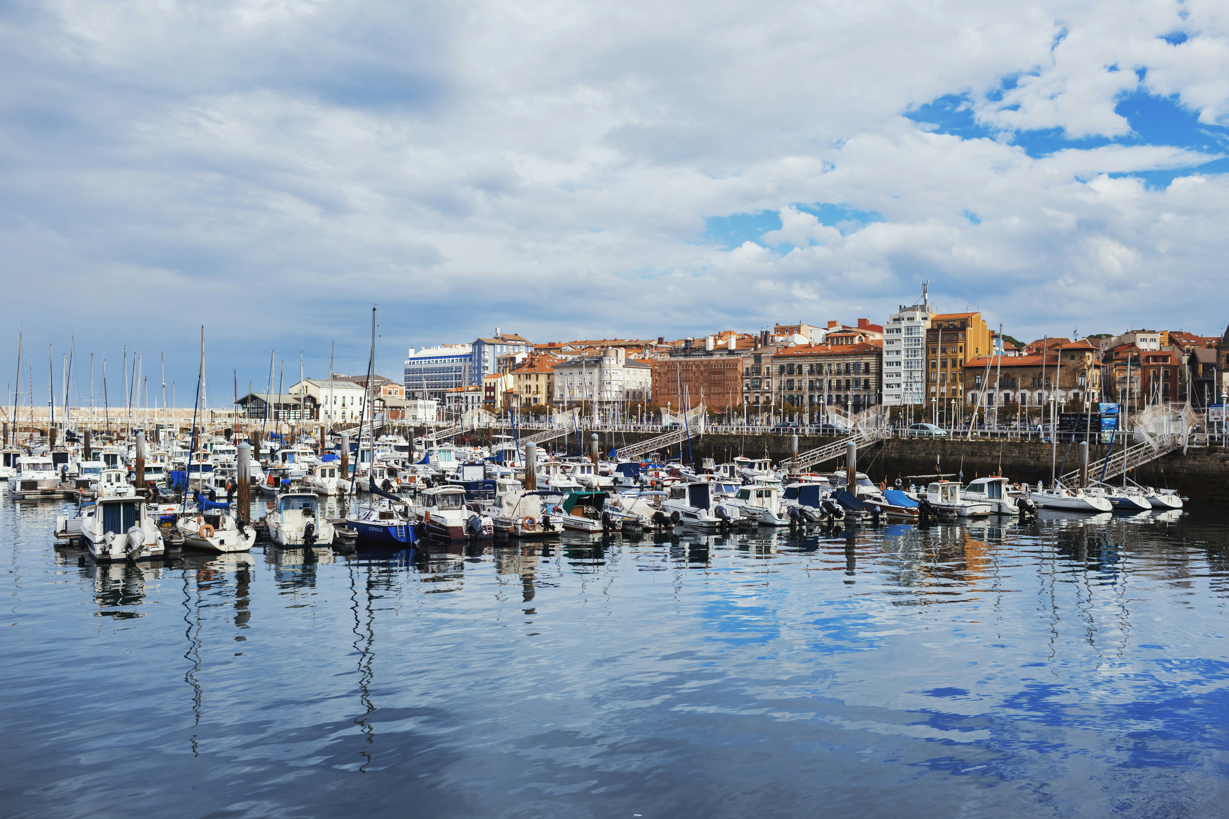 Boats docked in a harbor with city buildings behind. - Gijón