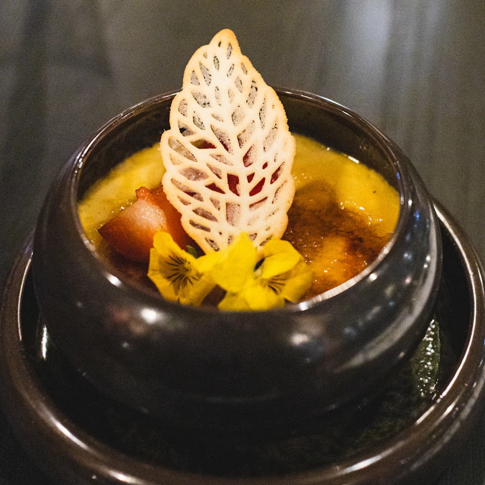 The image shows a small black bowl filled with a creamy yellow substance, possibly a dessert or sauce. The bowl is placed on a dark wooden table, and there are two additional bowls visible in the background - one larger than the other. A white leaf-shaped decoration adorns the top of the smaller bowl, adding an artistic touch to the presentation.

The main focus of the image is the small black bowl with its creamy yellow substance, which appears to be a dessert or sauce. The background features a dark wooden table and two additional bowls - one larger than the other.