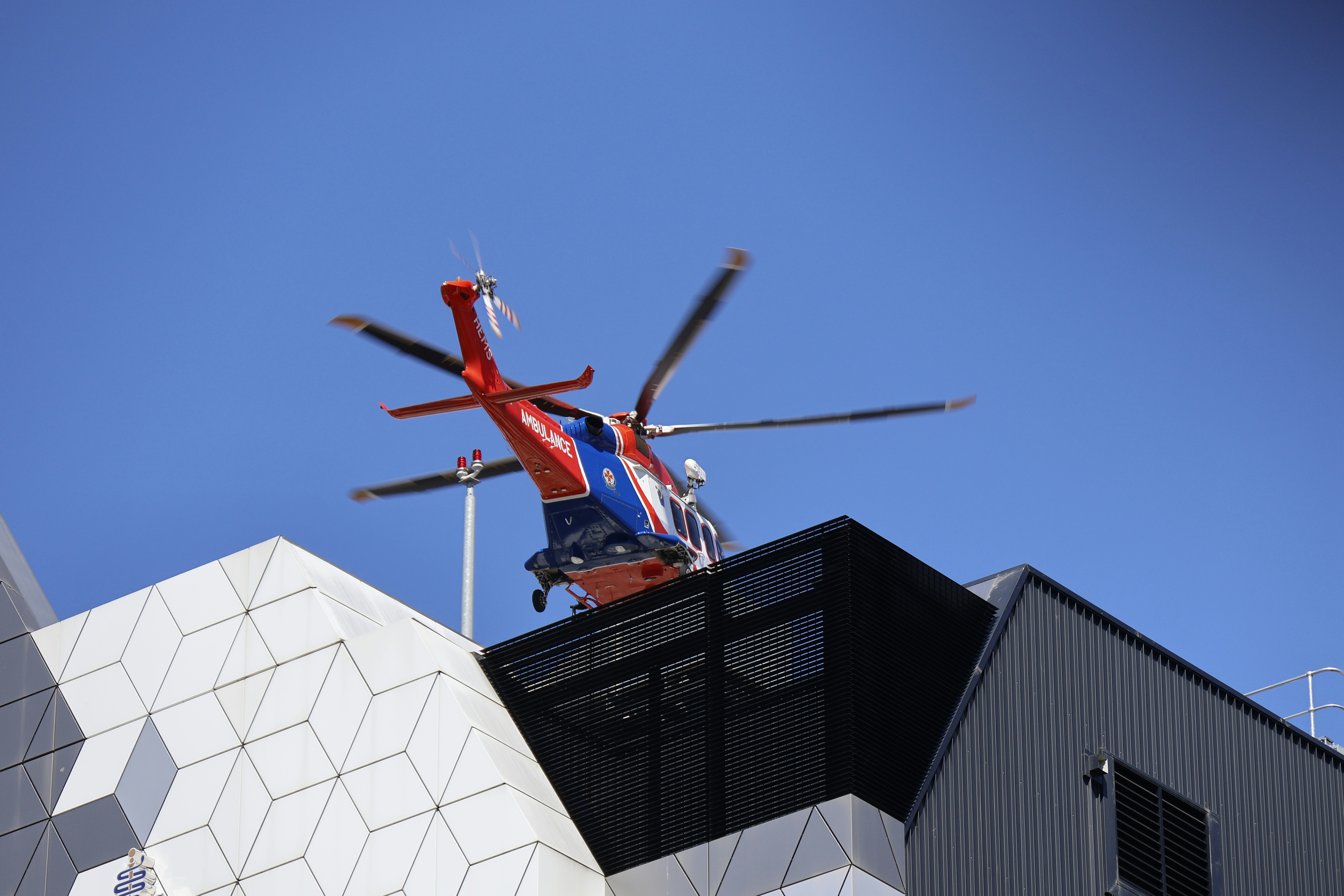 An emergency medical helicopter, red and blue, lands on a modern, geometric building helipad under a clear blue sky.