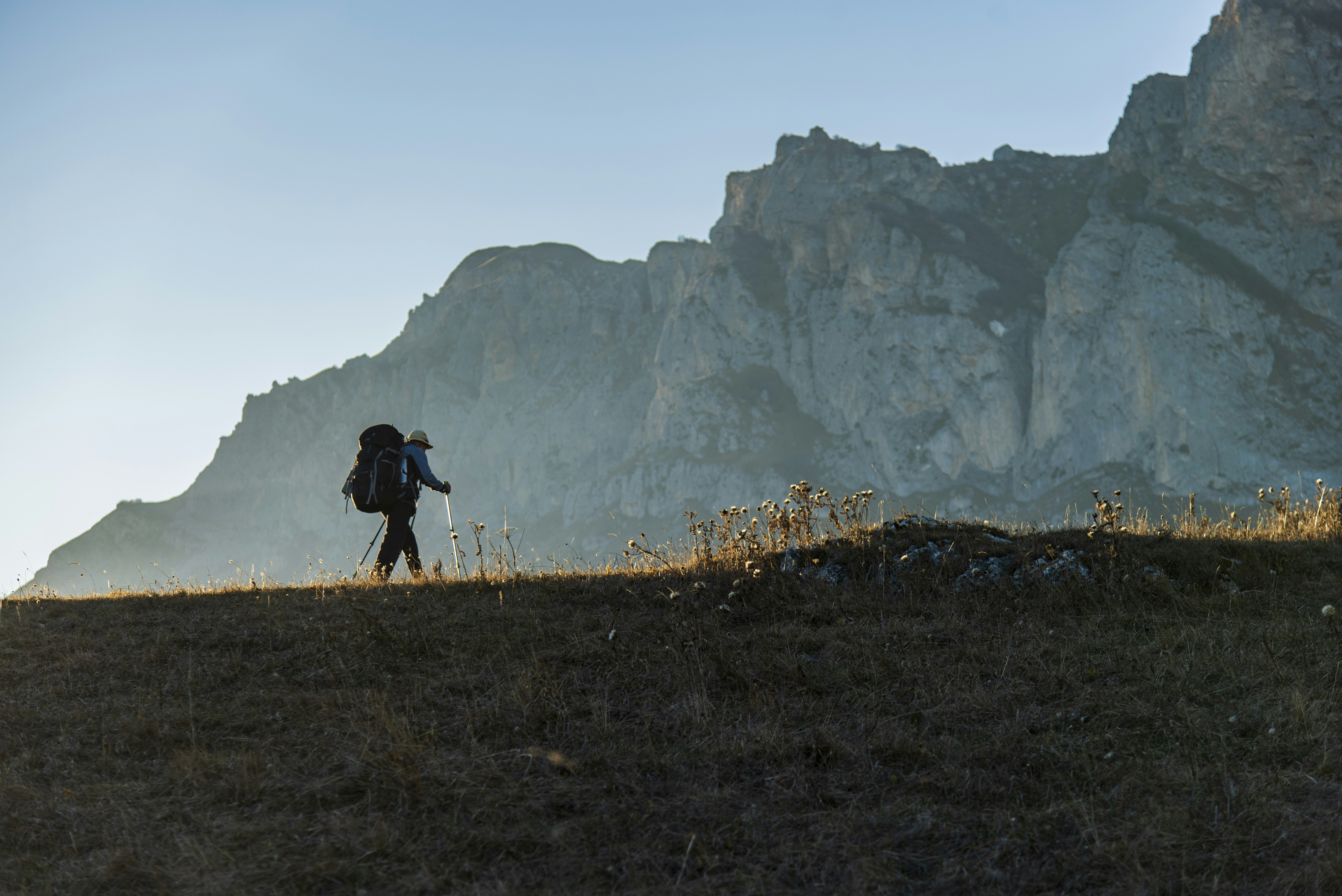 Hiker with backpack walks on a grassy ridge near mountains.