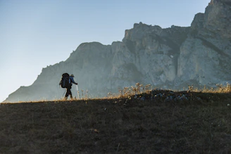 Hiker with backpack walks on a grassy ridge near mountains.