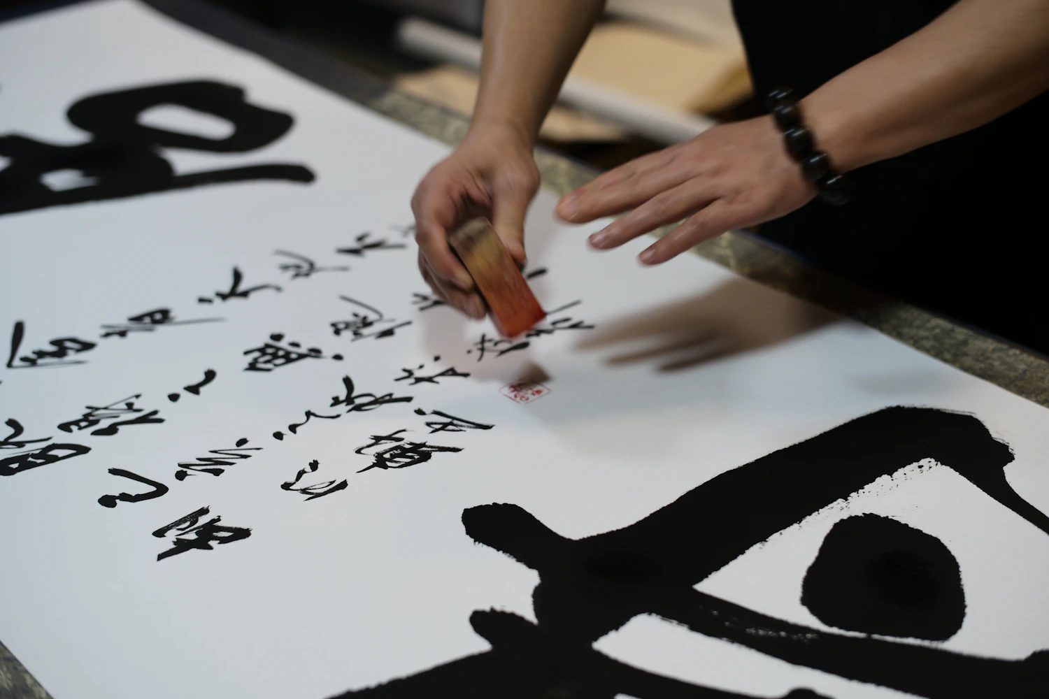 A hand pressing a brush into black ink strokes on a large white sheet