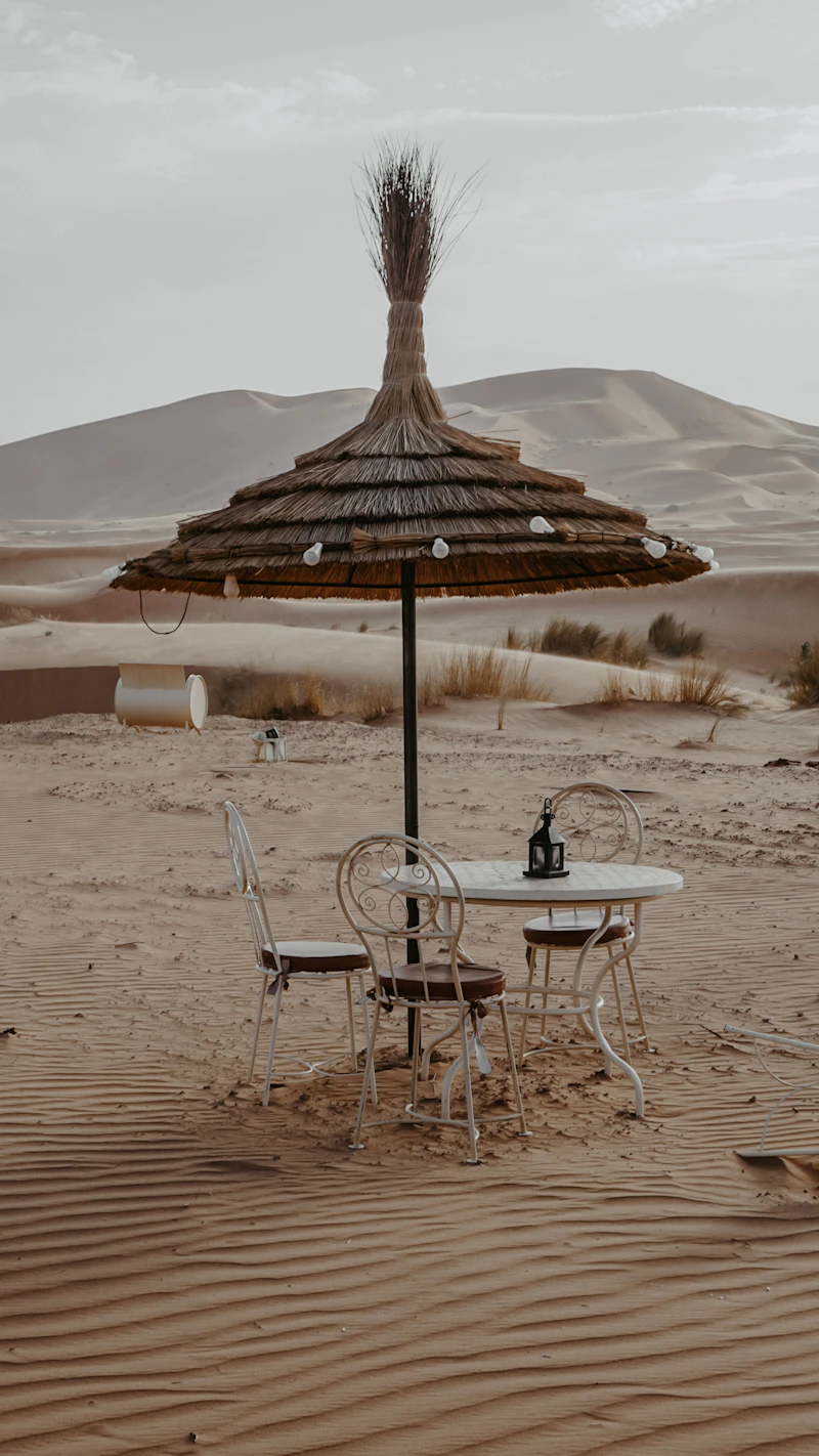 A shaded table and chairs under a straw parasol in the dunes