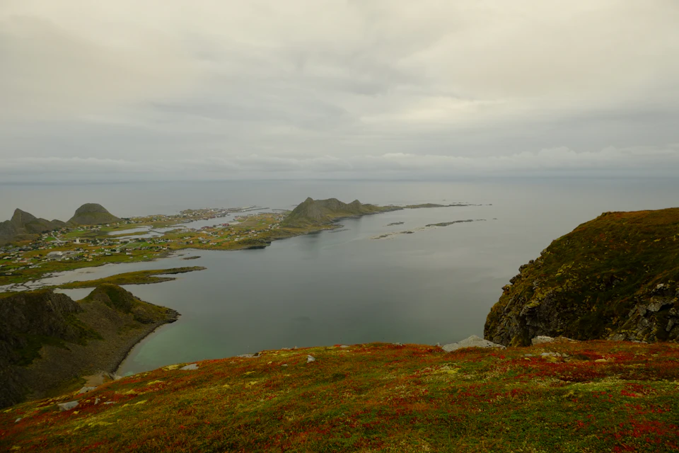 The image depicts a serene coastal landscape with a rocky cliff overlooking a deep blue sea. The sky above is overcast, casting a soft light on the scene below. On the left side of the image, there are several small islands scattered across the water, adding to the picturesque view. In the foreground, a grassy hill rises up, providing a sense of depth and perspective to the landscape.