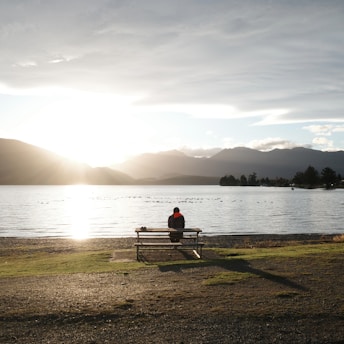Person sits on bench overlooking a tranquil lake at sunset.