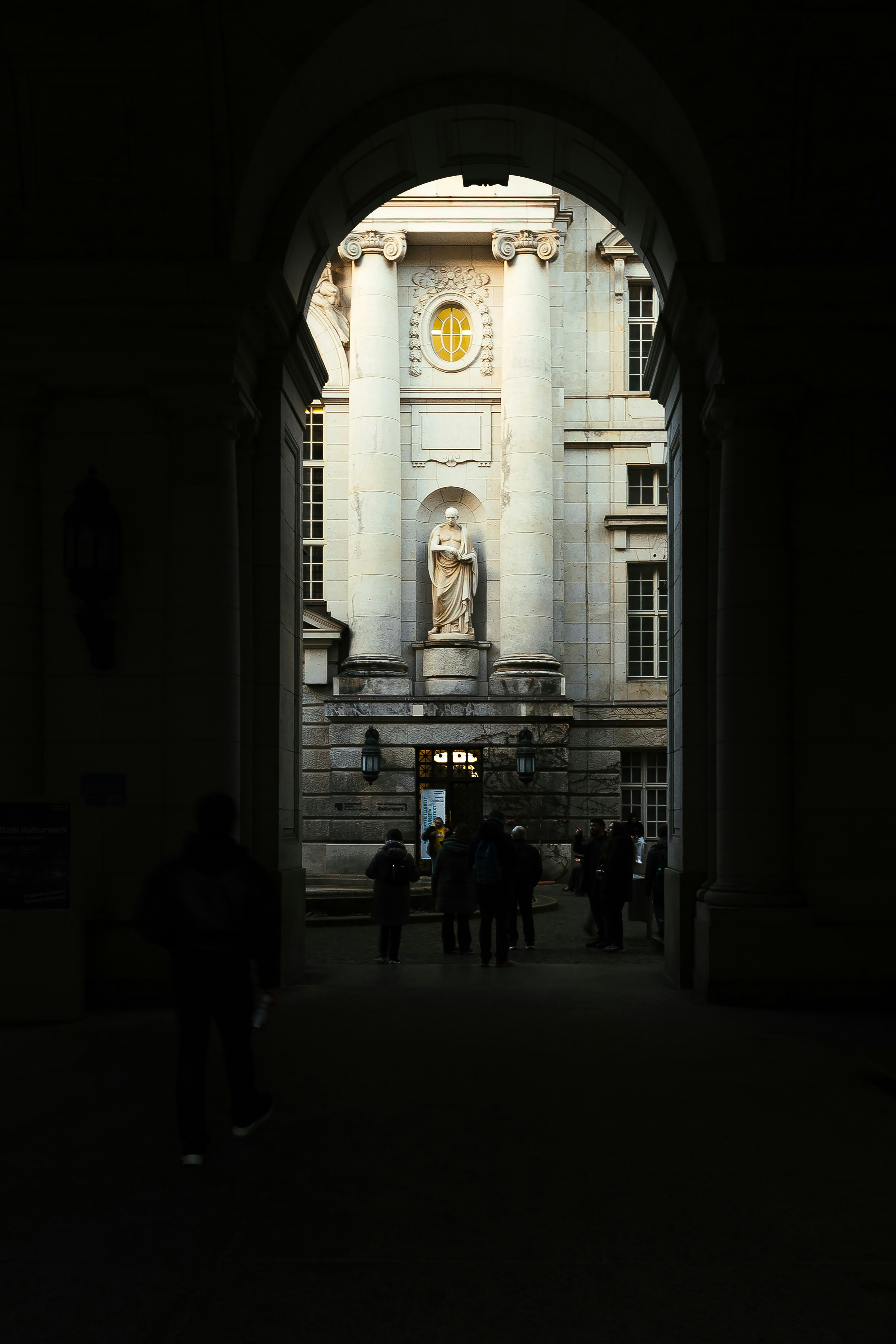 People walk through archway towards building with classical building.
