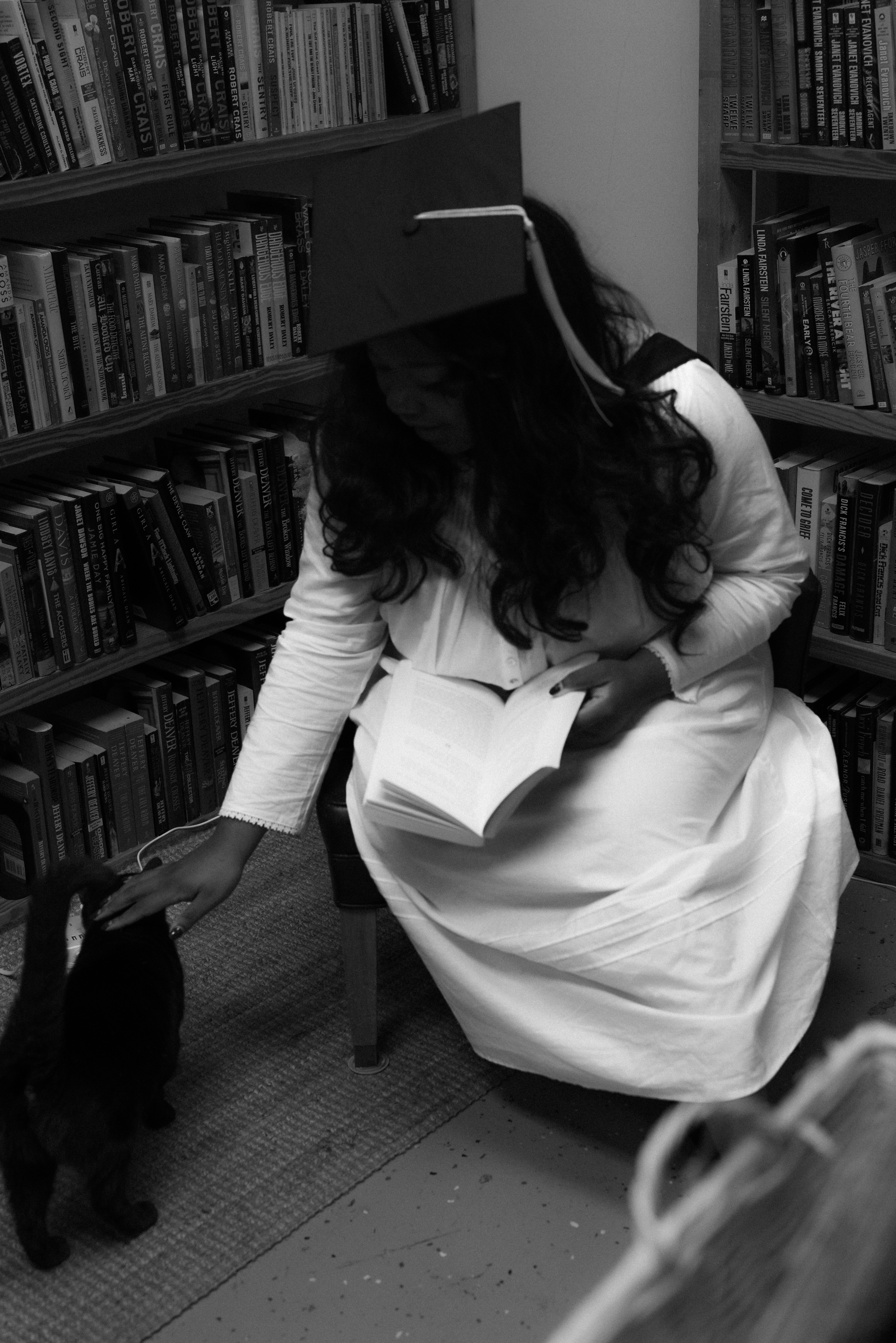 Woman in graduation cap petting black cat near bookshelves