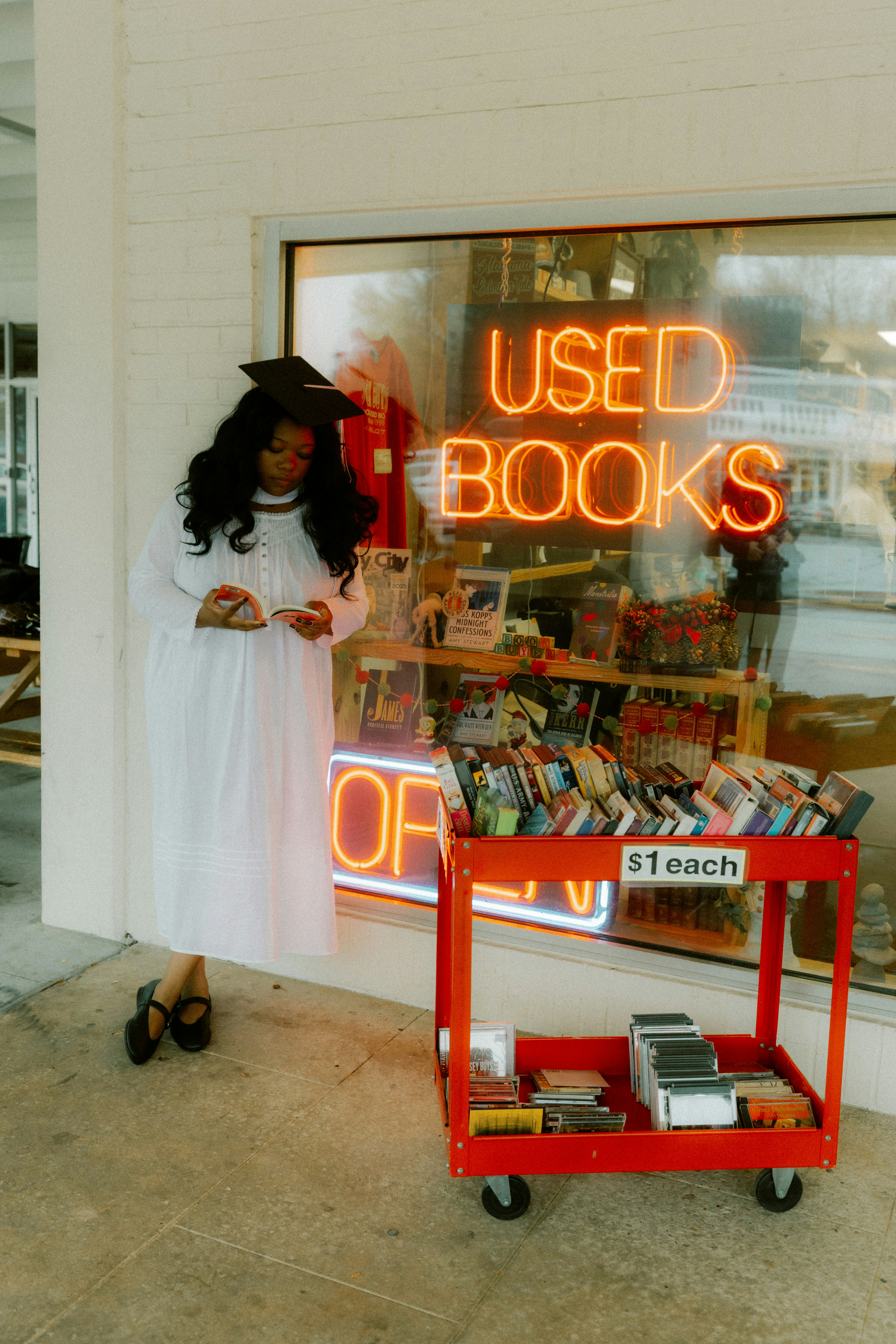 Woman in graduation gown reading book outside used bookstore