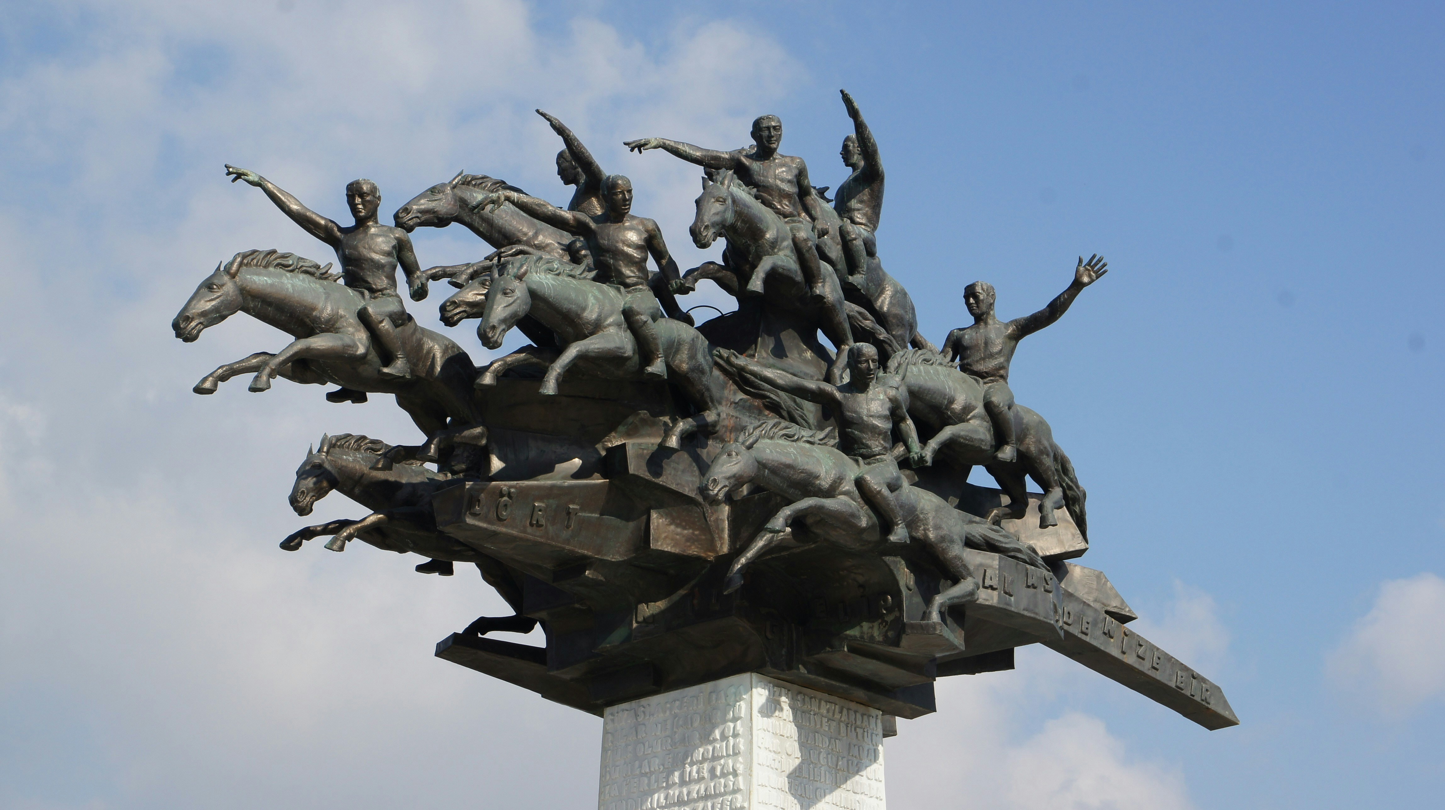 A close-up view shows the powerful expressions of riders and horses in the sculpture. The bronze figures appear energetic and full of motion. Details of muscles, gestures and dynamic lines are clearly visible. The bright sky behind them enhances the dramatic effect. The artwork expresses strength and collective determination.