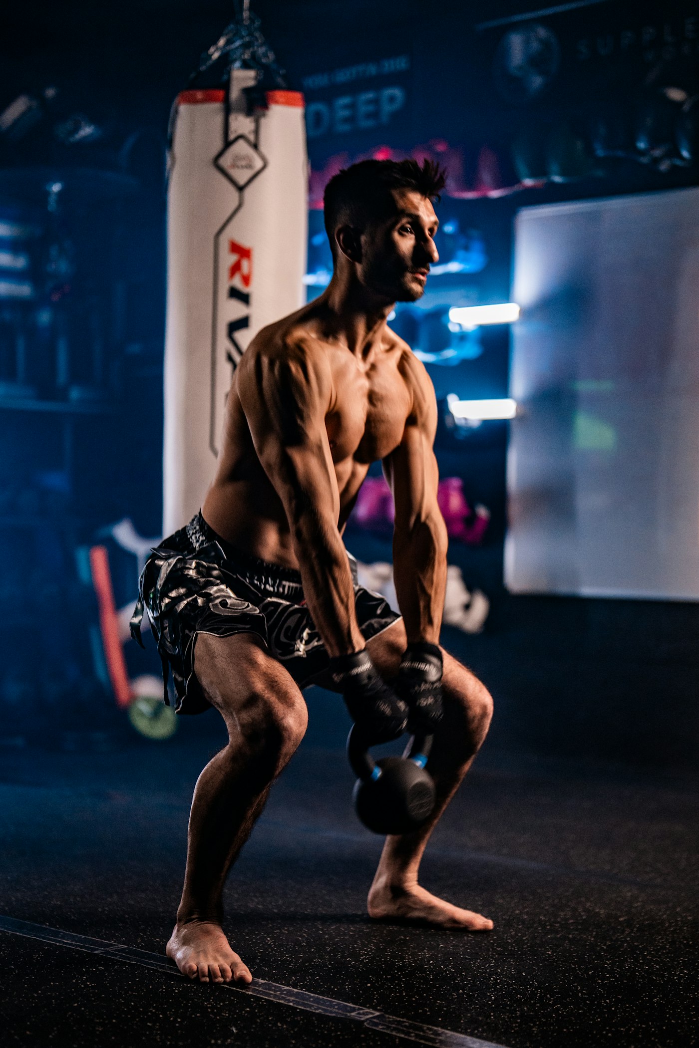 A barefoot man lifting a kettlebell beside a punching bag in a blue-lit gym