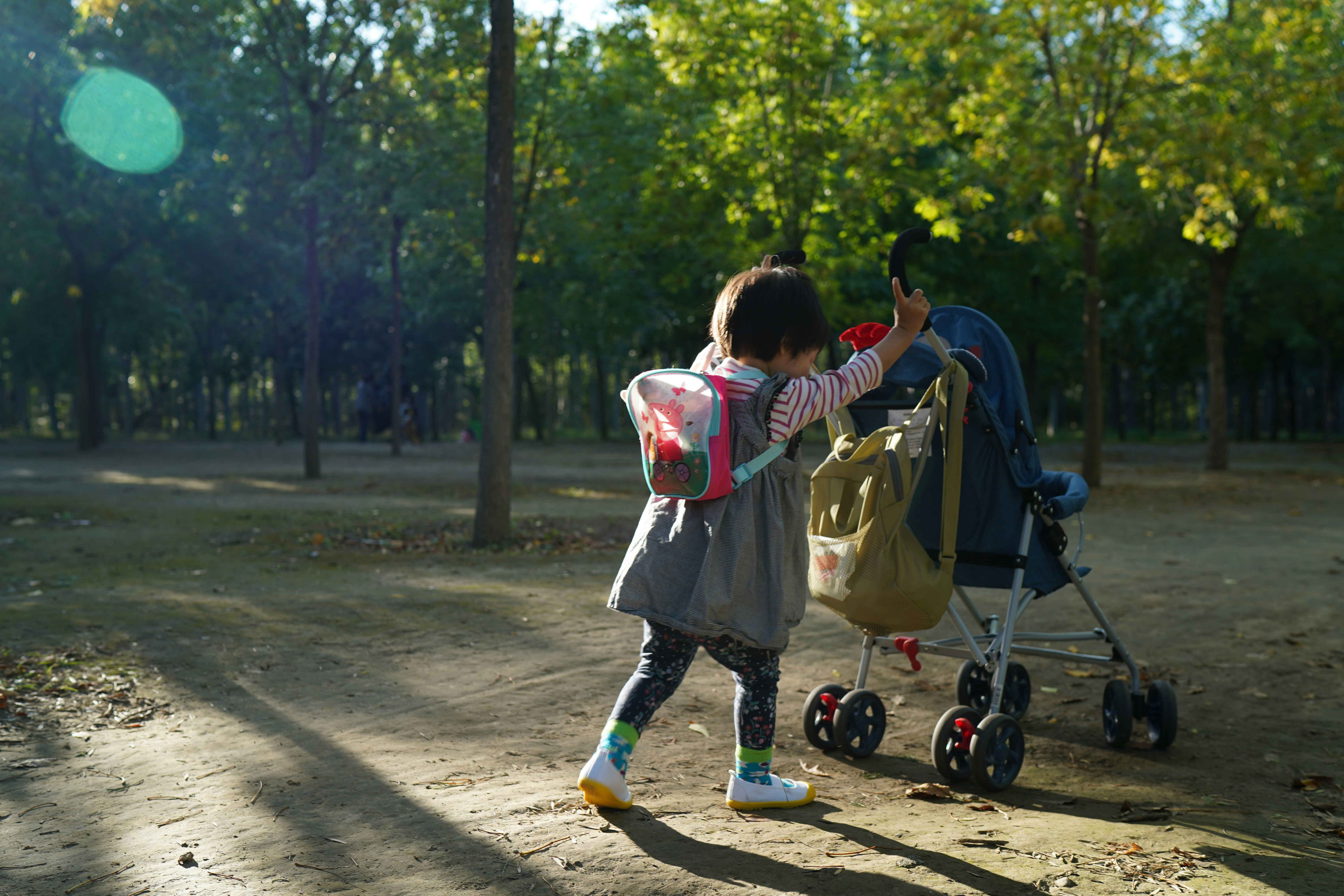 Child with backpack reaches for stroller in park.