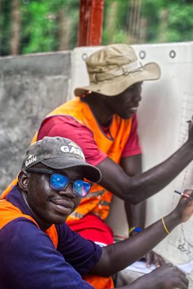 Two men wearing hats and vests writing on paper.
