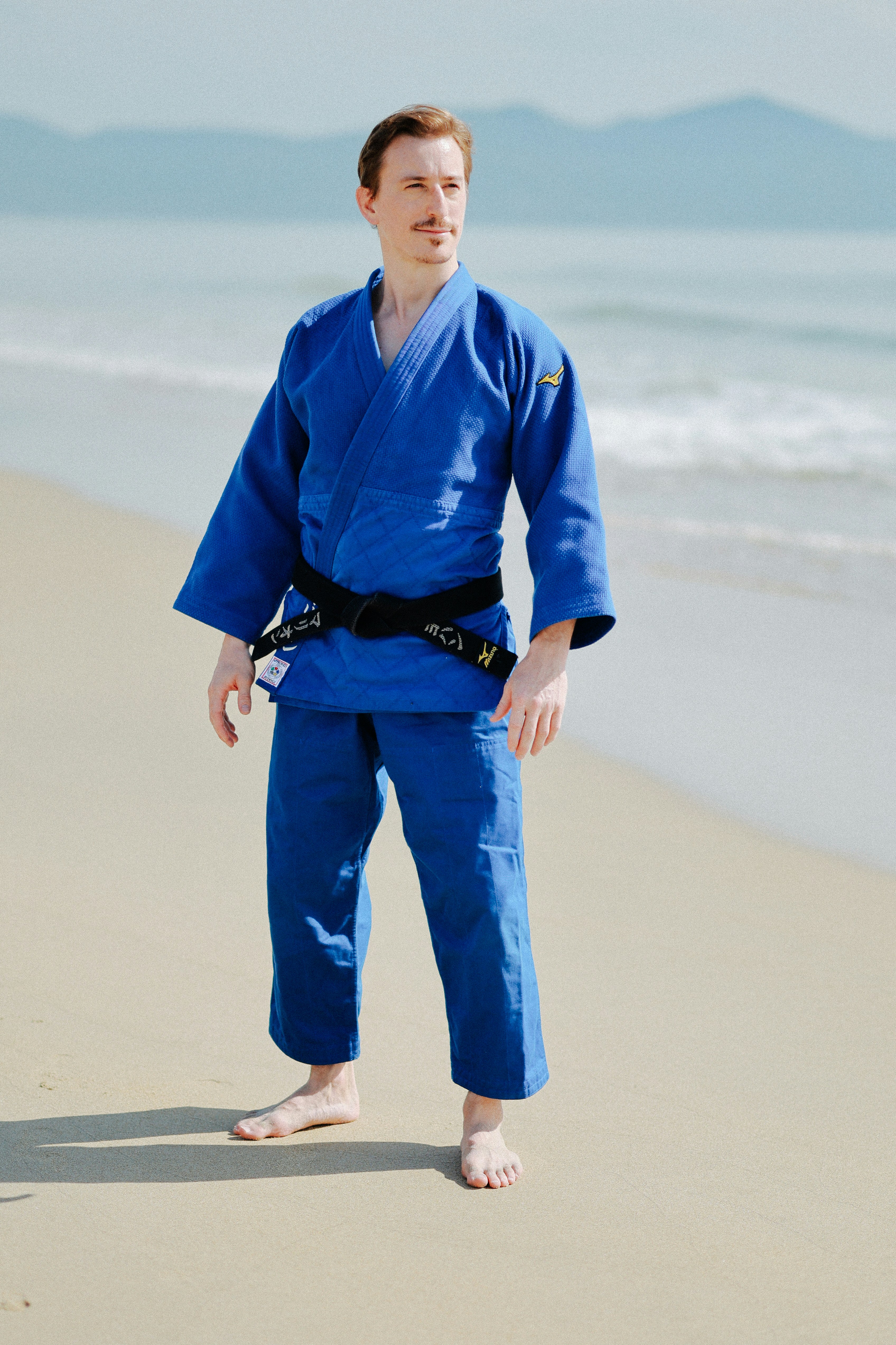 Man in blue judo gi on a sandy beach.