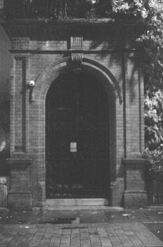 A grainy black-and-white photograph of a brick arched doorway with a white security camera on the left jamb