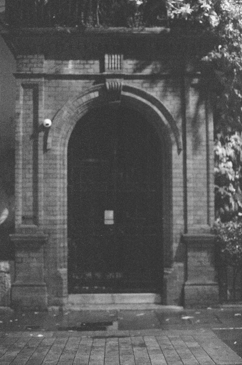 A grainy black-and-white photograph of a brick arched doorway with a white security camera on the left jamb