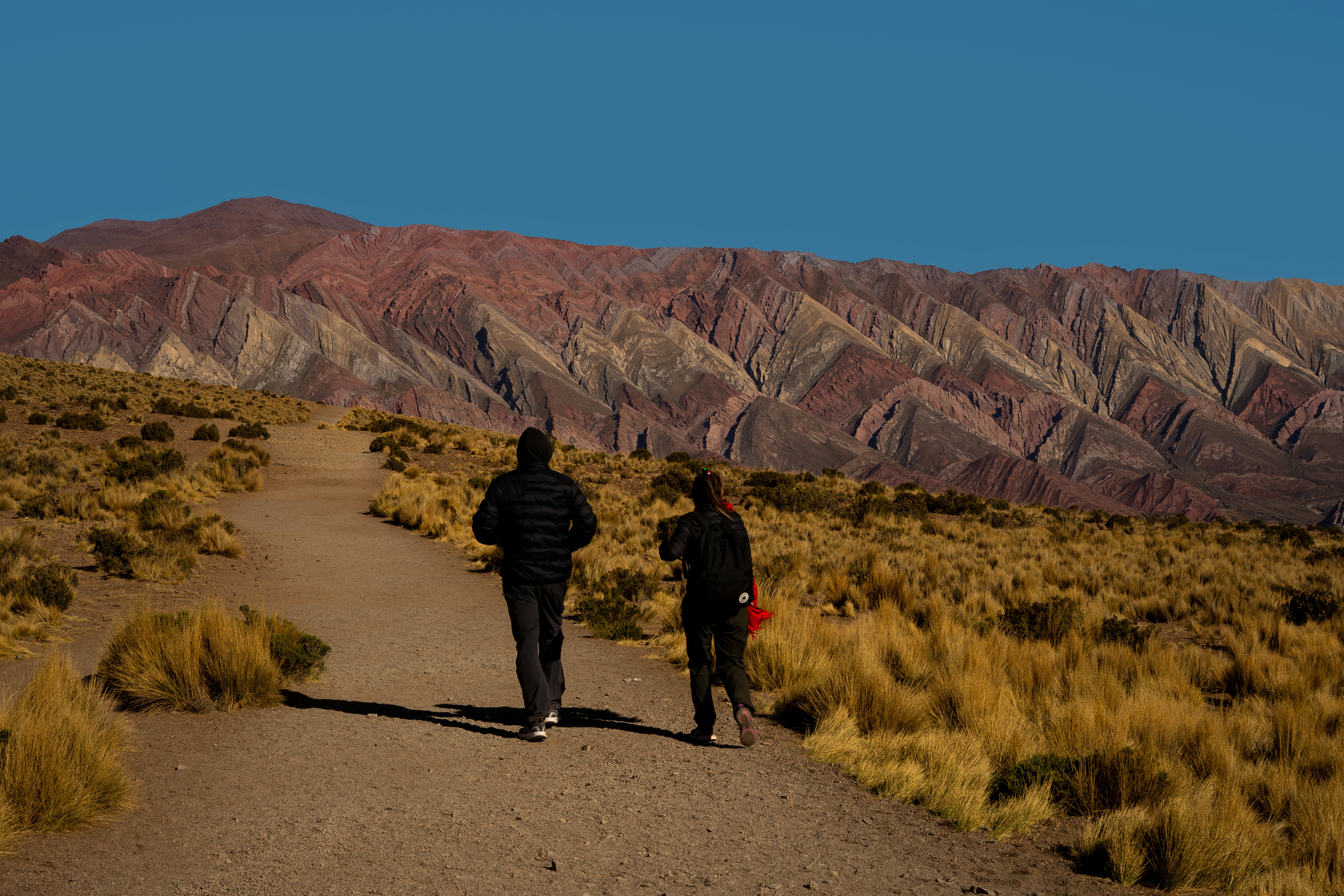 Dos personas caminan por un sendero hacia montañas coloridas.