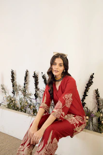 Young woman in a red embroidered outfit sits near plants.