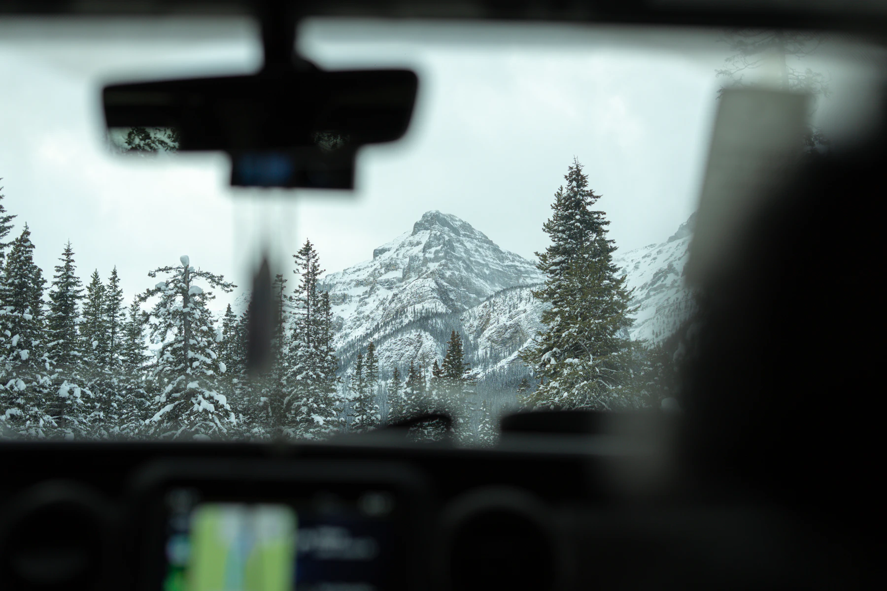 A snow-covered peak and snow-laden spruce trees seen through a car windshield, the dashboard and mirror out of focus in front