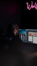 Man with braids working on computer with neon sign.