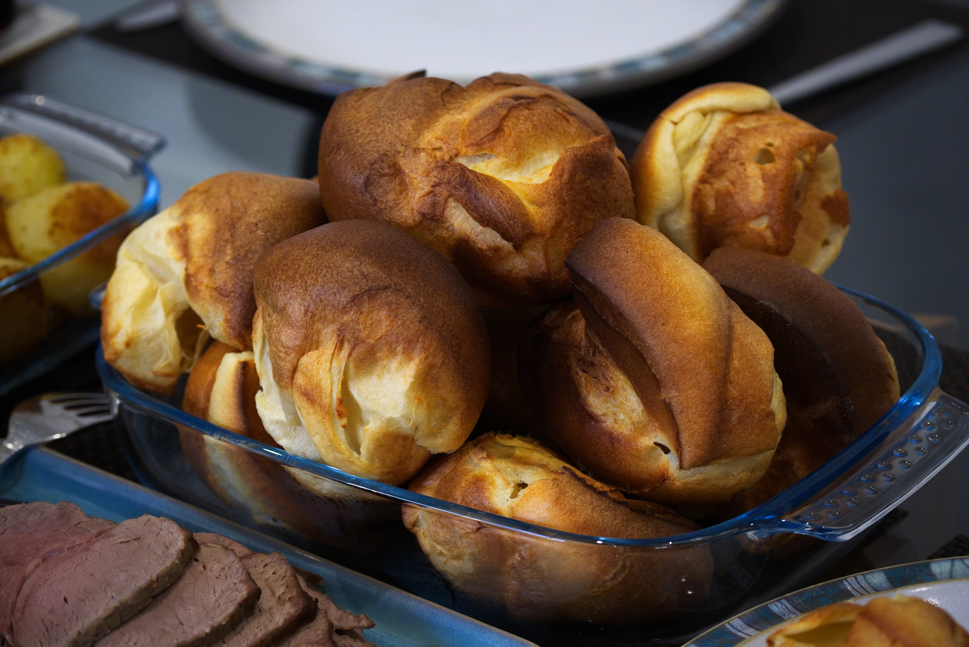 A glass dish piled with golden, crisply risen Yorkshire puddings, with a plate of sliced roast beef alongside — the signature accompaniment to a Yorkshire Sunday roast