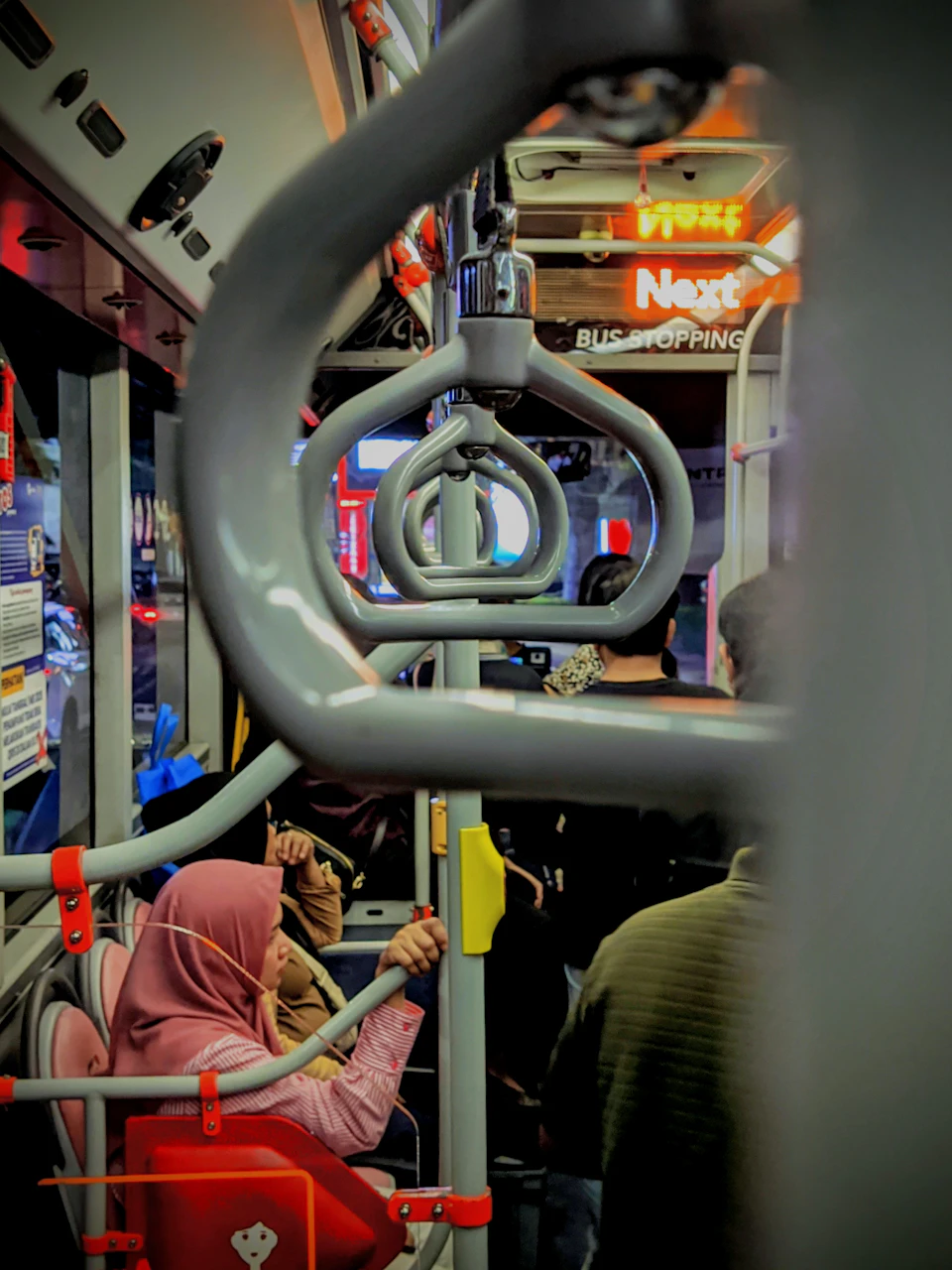 The image shows a bus interior with passengers seated and standing, including two individuals wearing pink headscarves - one person is sitting down while another stands up holding onto a pole. The bus has a gray exterior and red accents on its doors and windows, creating a vibrant contrast against the neutral background of the interior. A sign reading "Next Bus Shopping" can be seen in the background, indicating that this is likely a transit hub or bus station where passengers can board buses for shopping or other destinations.

The image also contains text elements such as "First", "Second", and "Third", which could potentially be part of a map or schedule displayed on the bus.