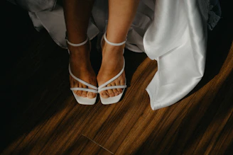 Bride's feet in white strappy heels on wooden floor.