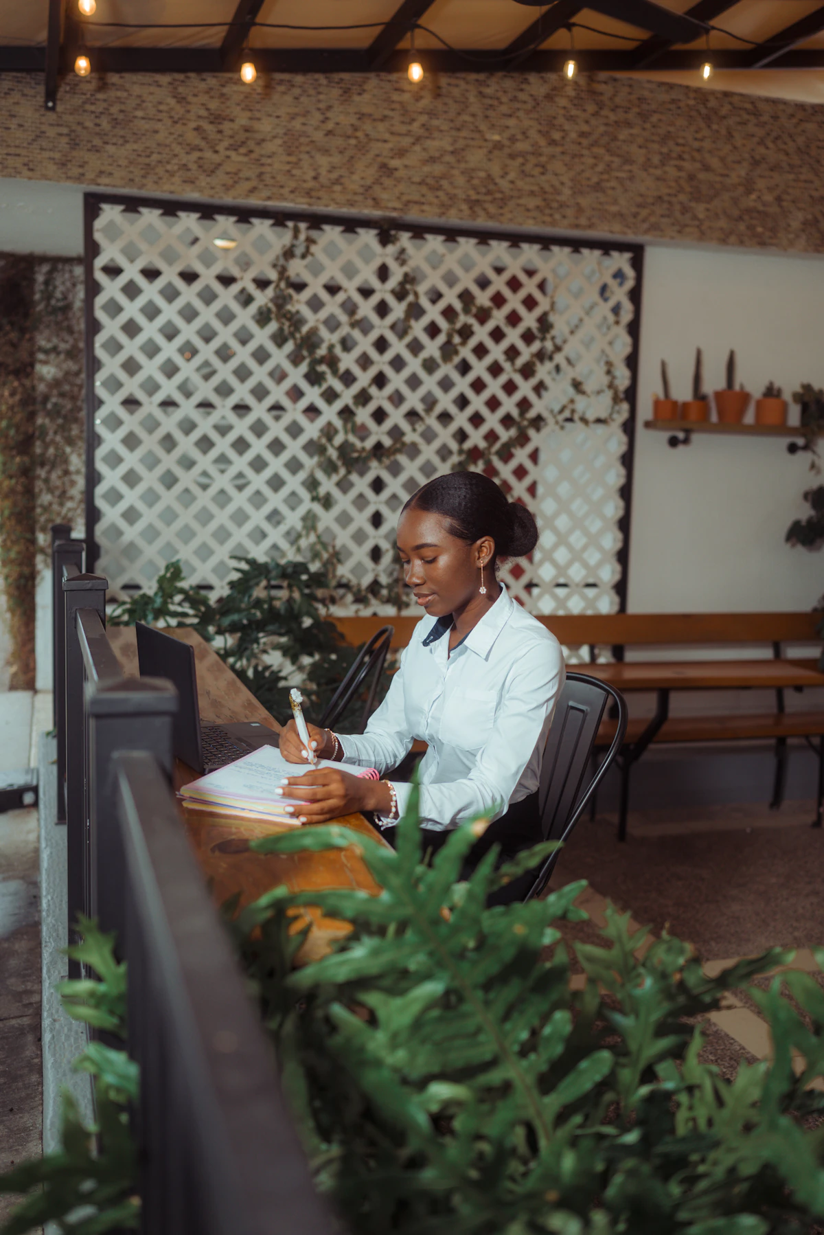 Une femme en chemise blanche écrit sur des feuilles à côté d'un ordinateur portable, installée à une table entourée de plantes