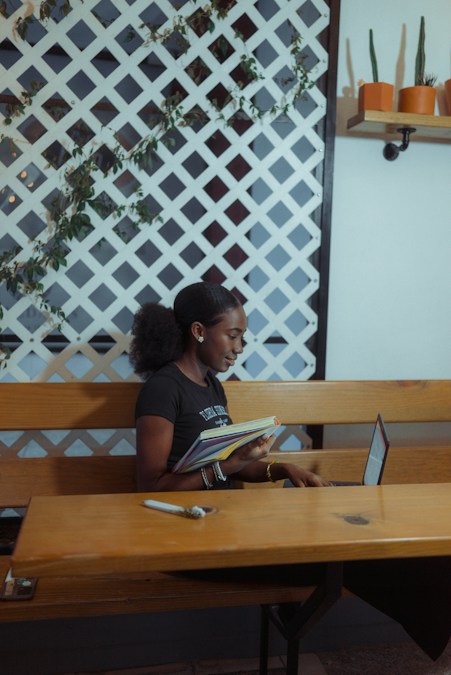 Une étudiante feuillette un cahier devant son ordinateur portable, assise à une table en bois