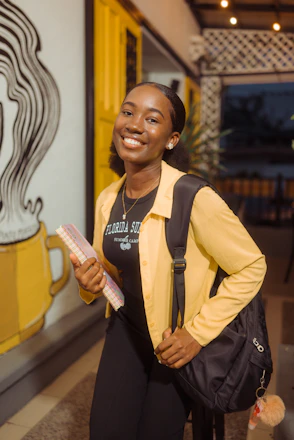 A smiling woman in a yellow shirt and black backpack holding a notebook