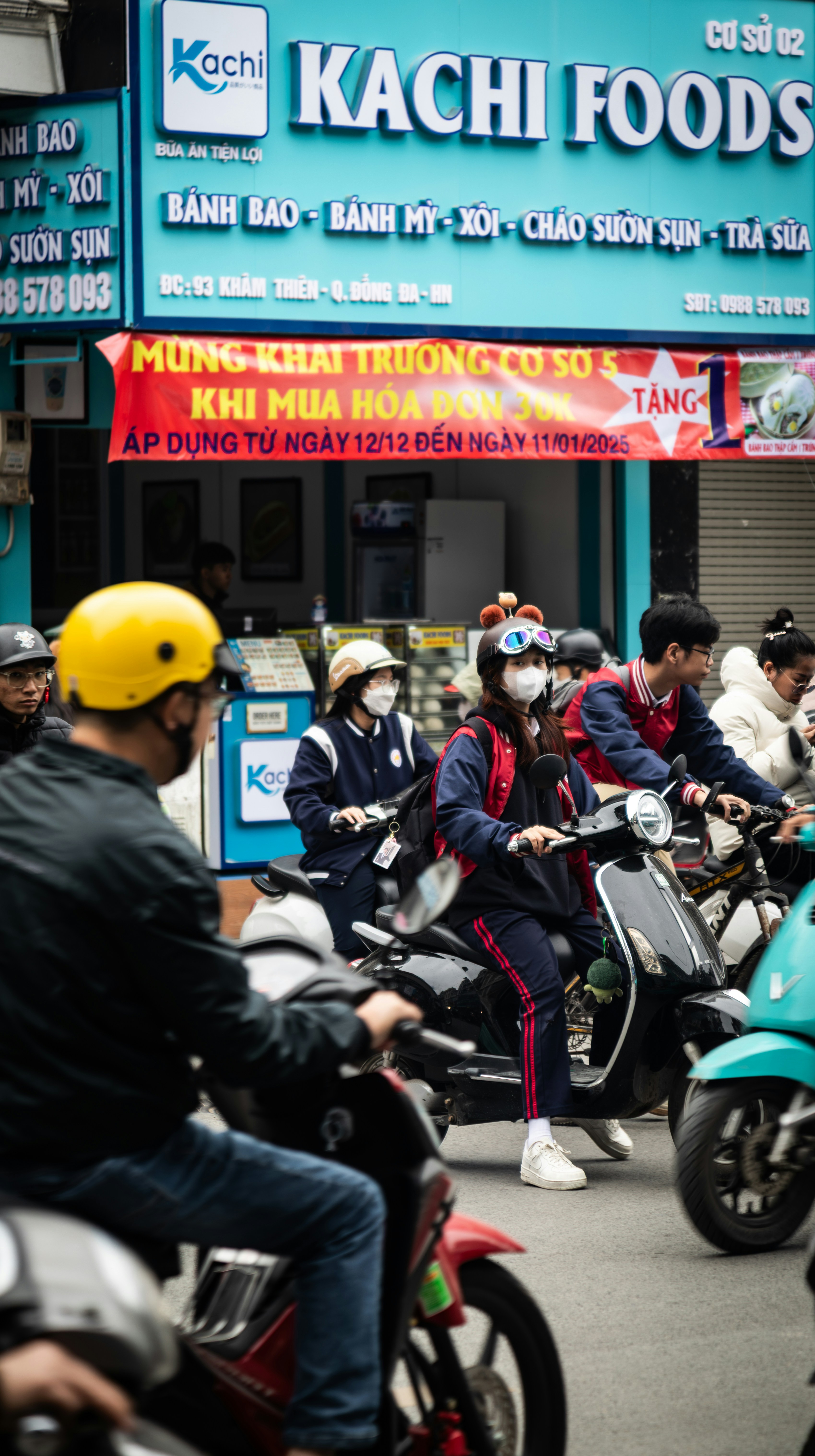 People wearing masks ride scooters past a kachi foods store.