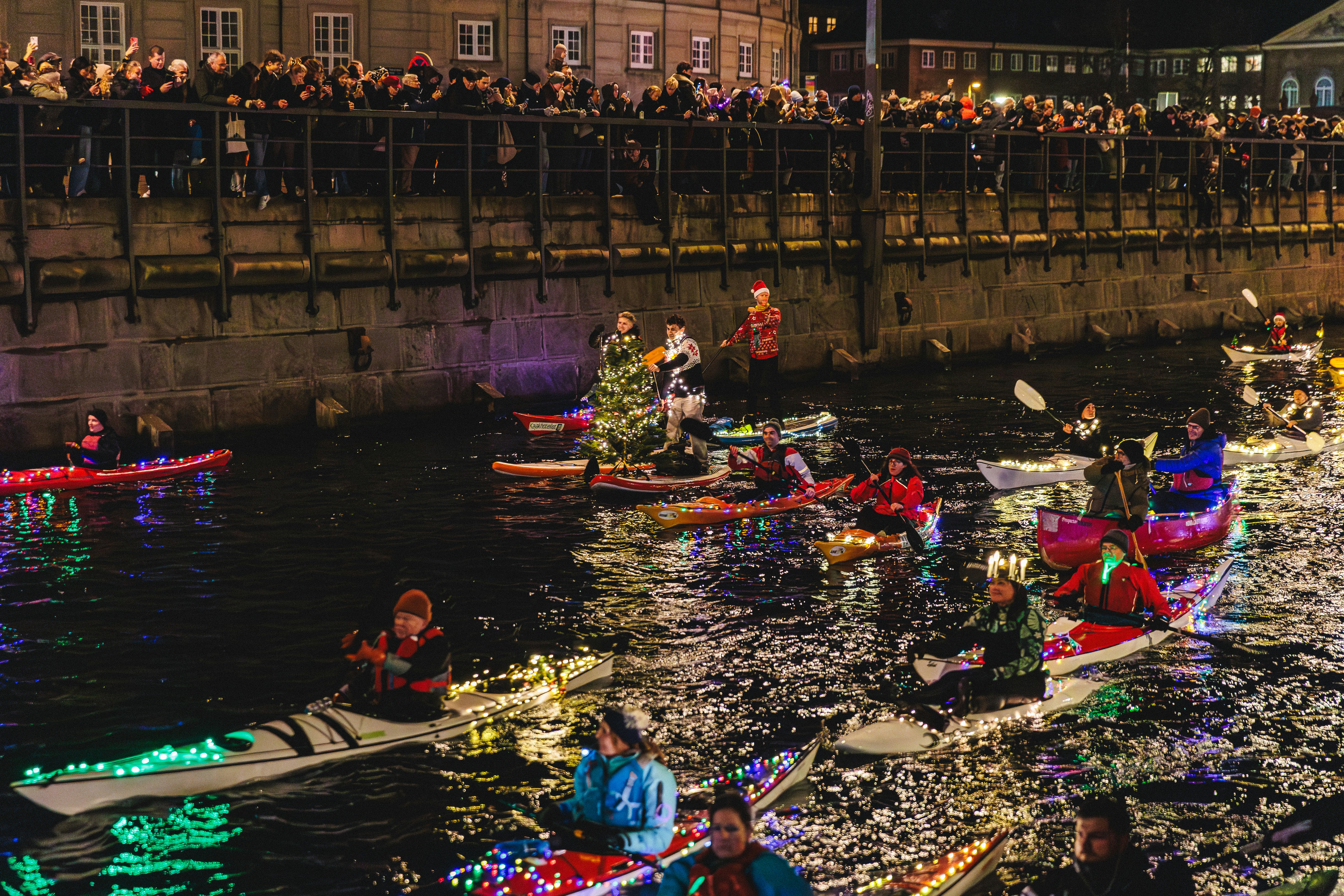 People in illuminated kayaks parade on a canal at night.