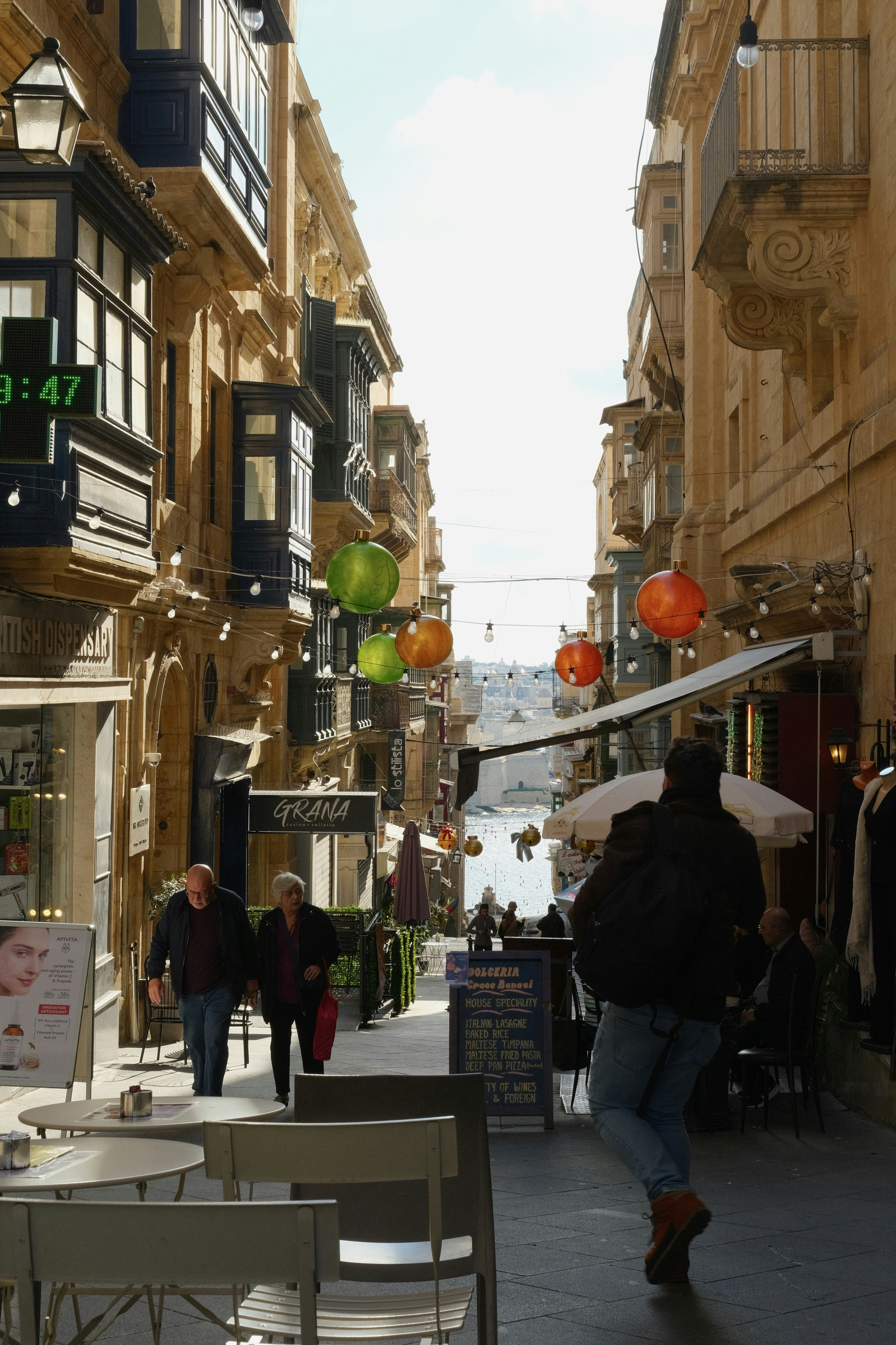 People walk down a narrow street with shops and cafes.