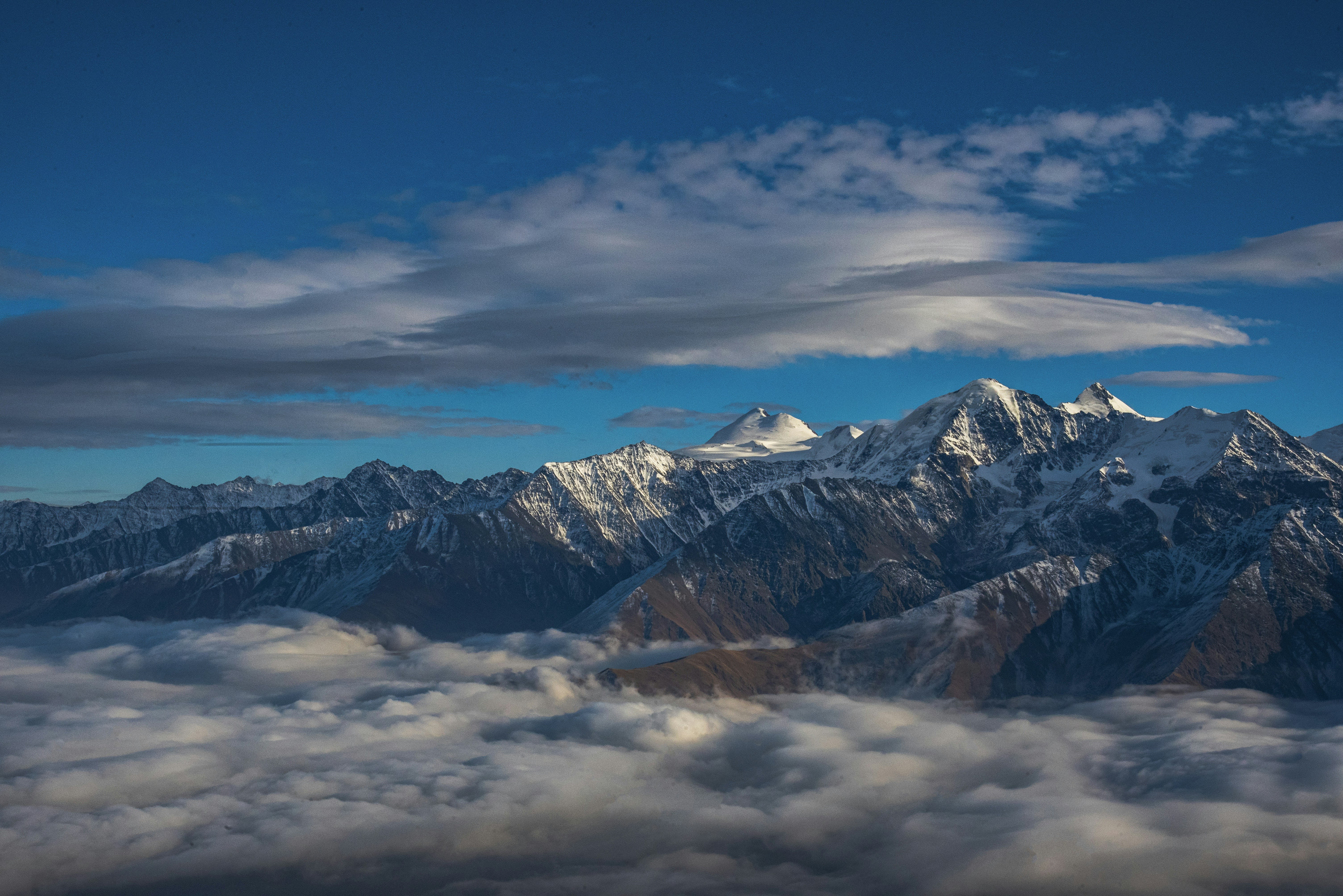 Snow-capped mountains rise above clouds under blue sky