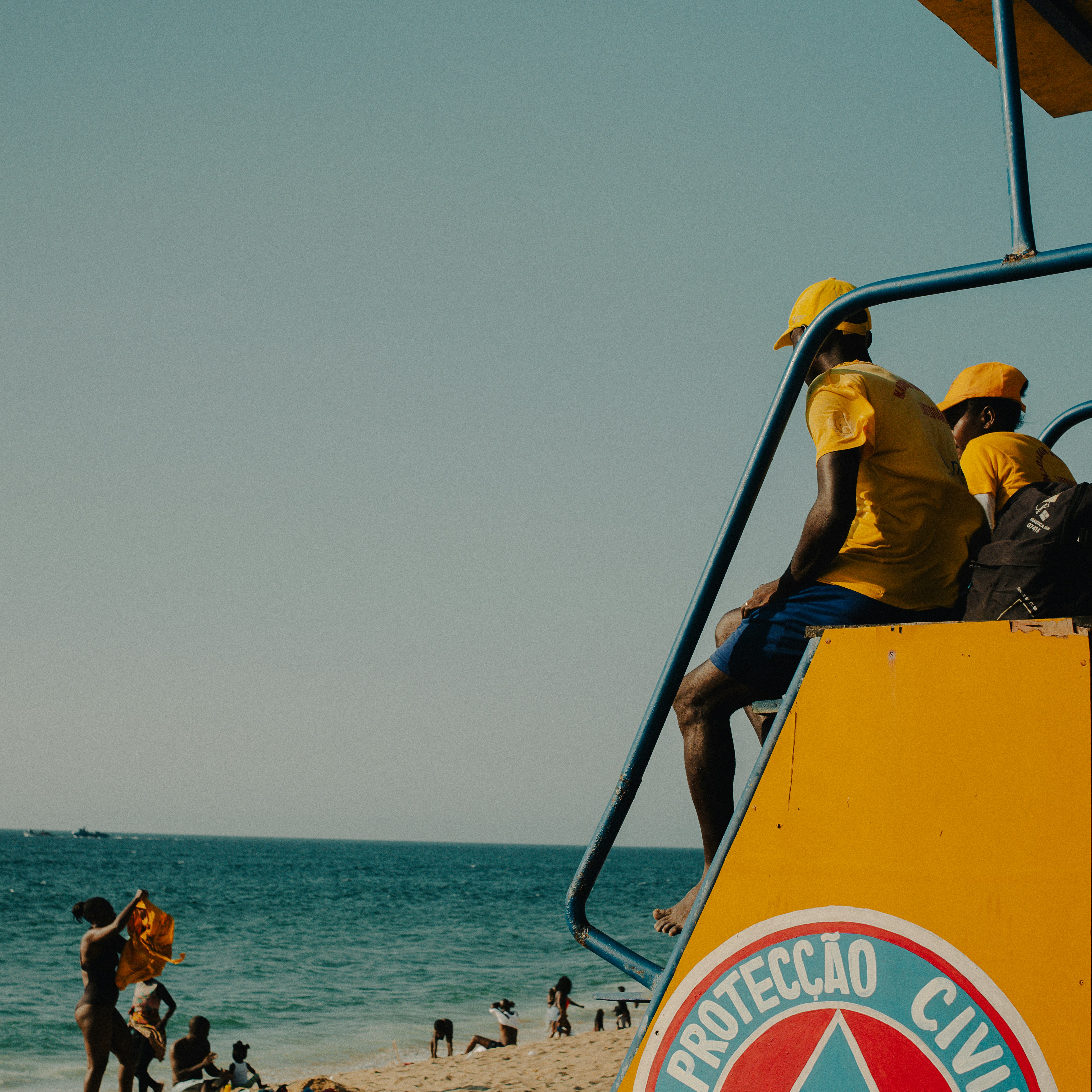 Lifeguards in yellow shirts watch beachgoers from tower.