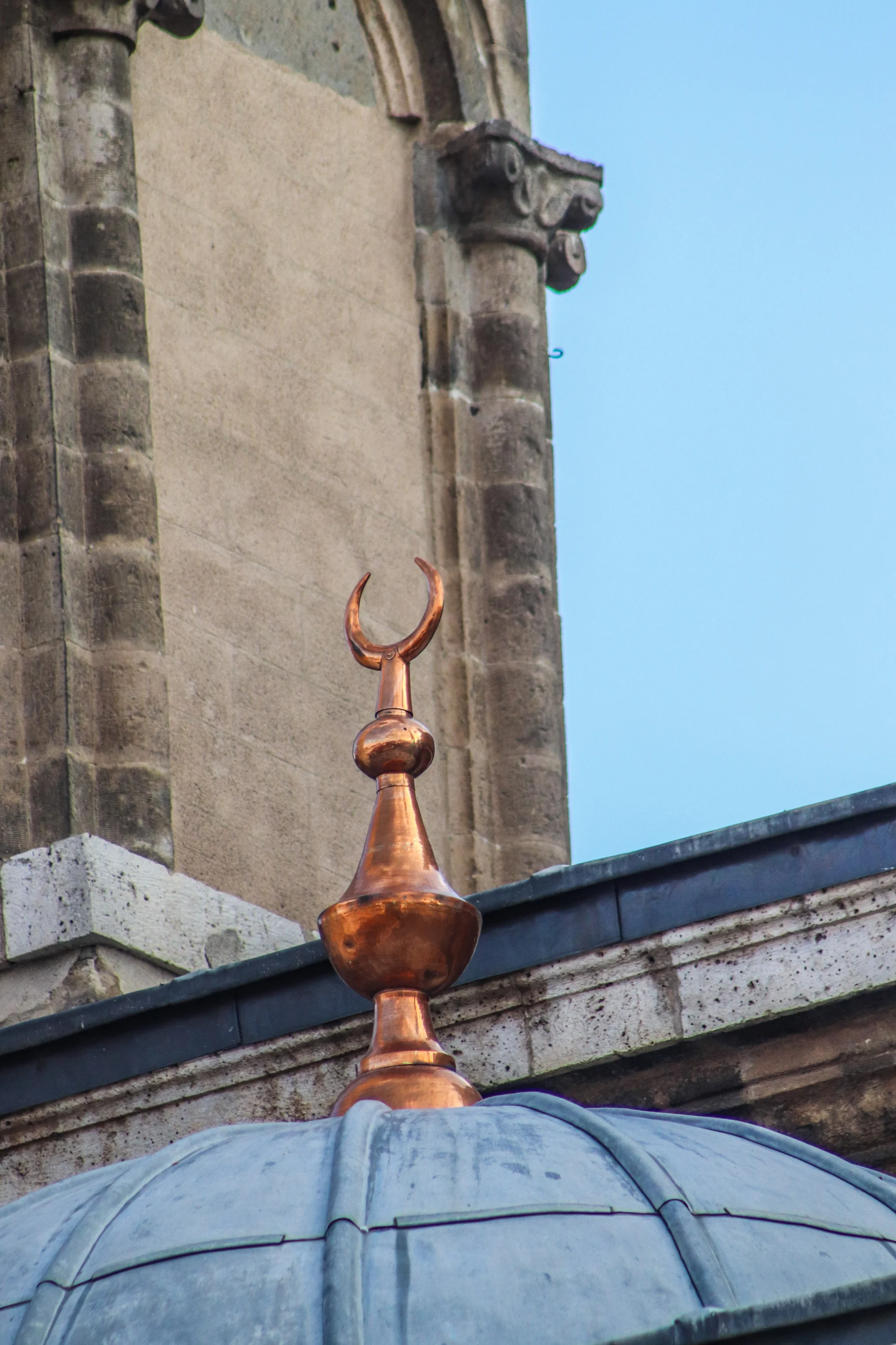A copper crescent finial on top of a grey mosque dome, with a stone minaret and blue sky behind it