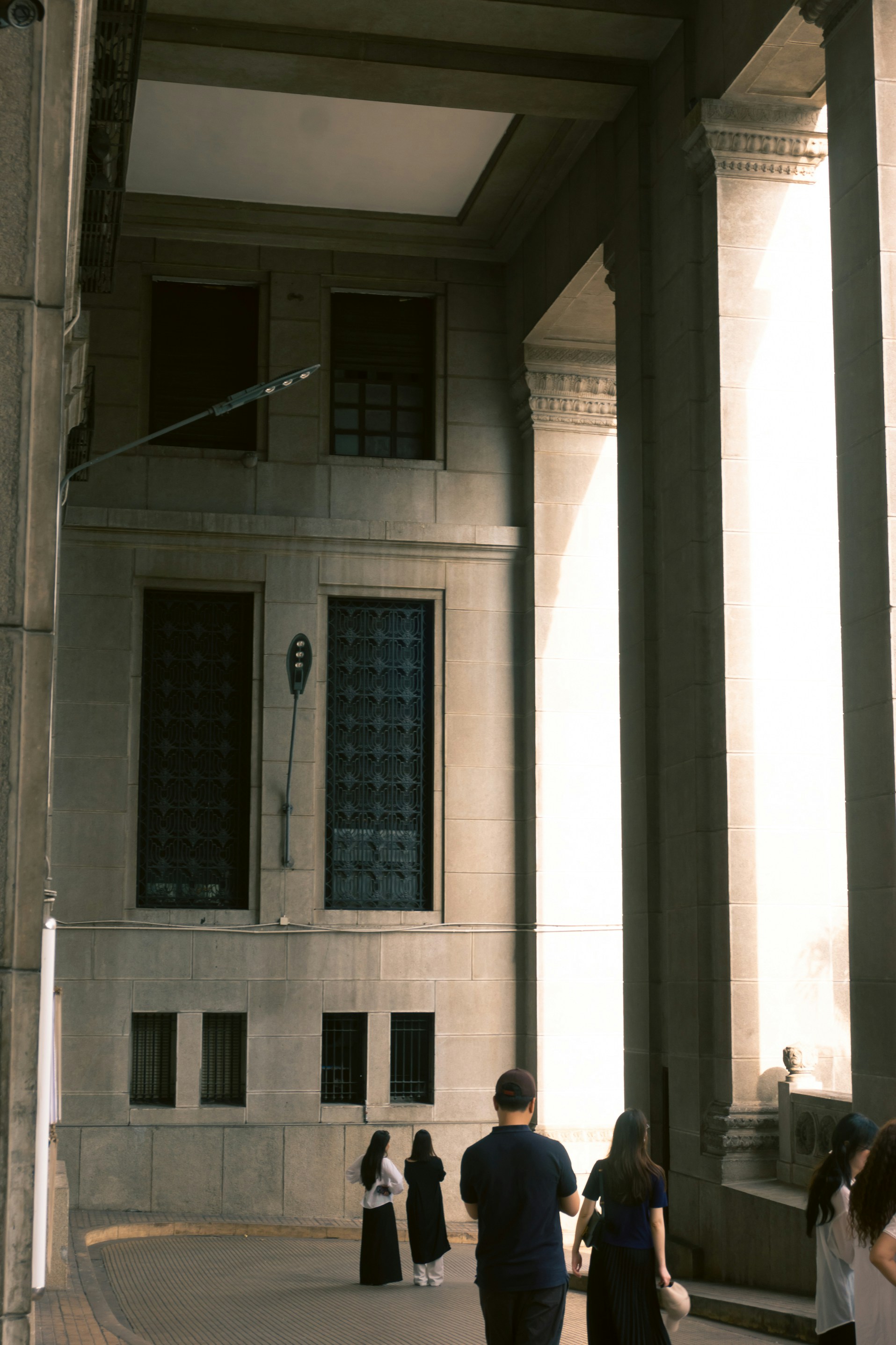 People walk through a grand stone colonnade with windows