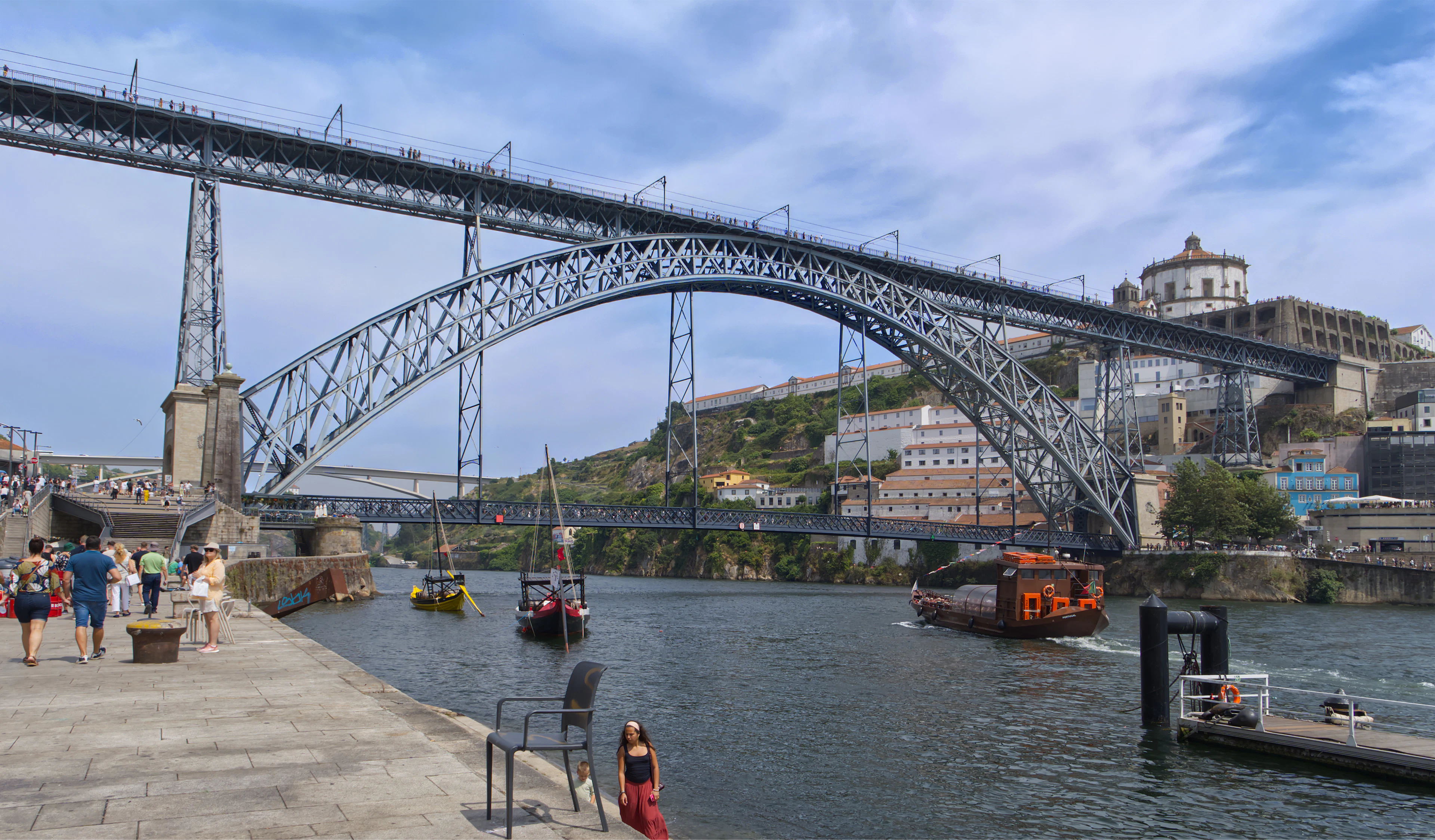 The iron arch of the Dom Luis I Bridge spanning the Douro, seen from the quayside with traditional rabelo boats moored below