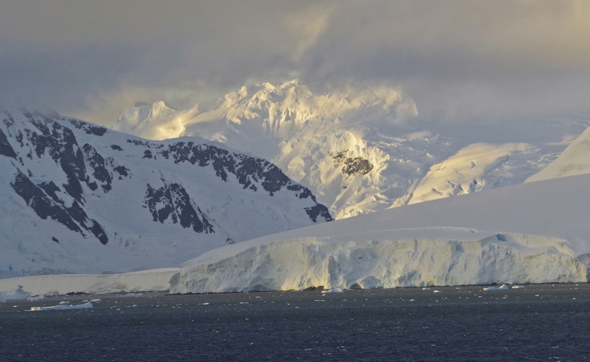 Snow-covered mountains under low cloud with a glacier ice cliff meeting dark open sea, warm low light catching the highest peaks and small pieces of floating ice in the foreground