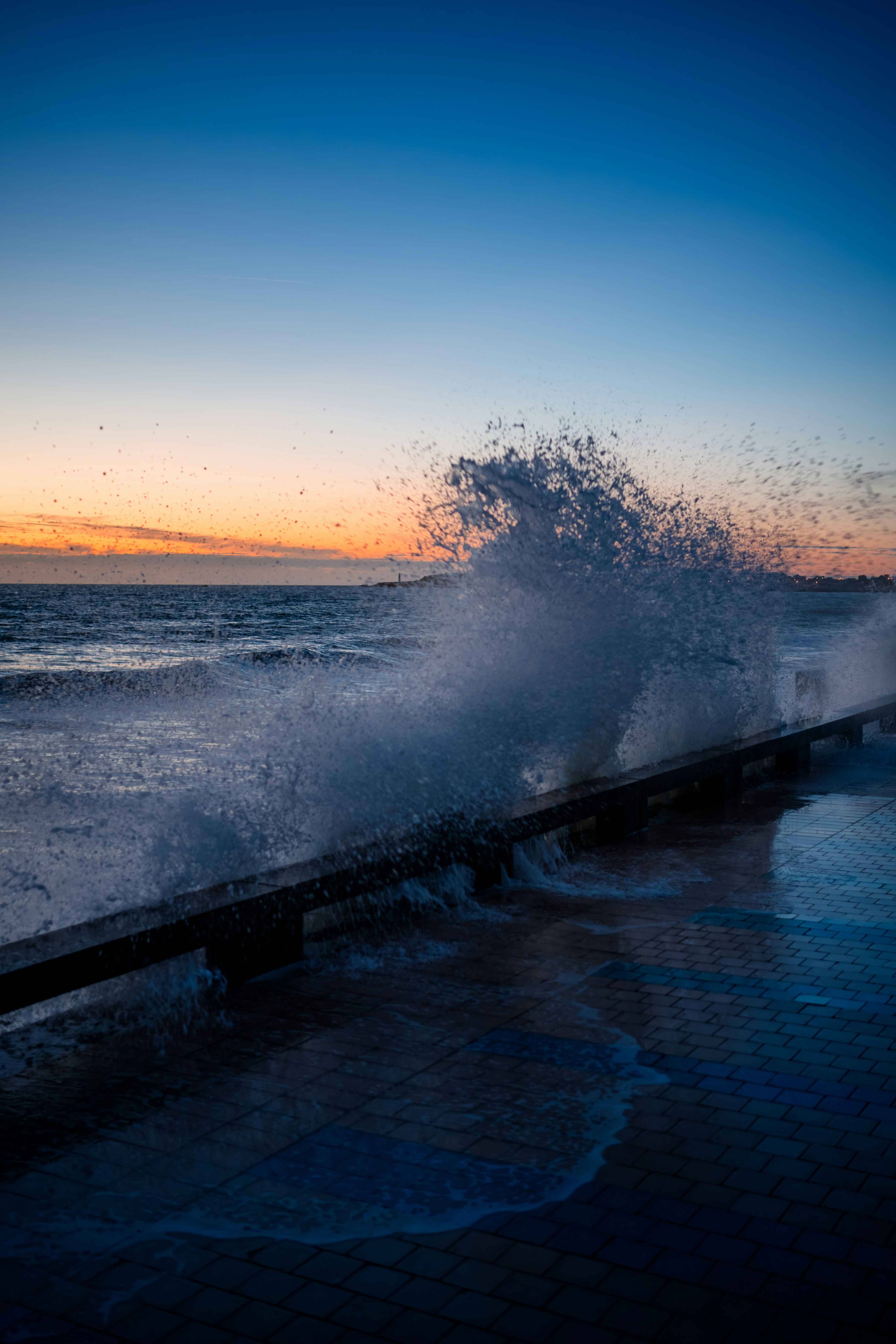 A large wave crashes against a wooden pier at sunset.