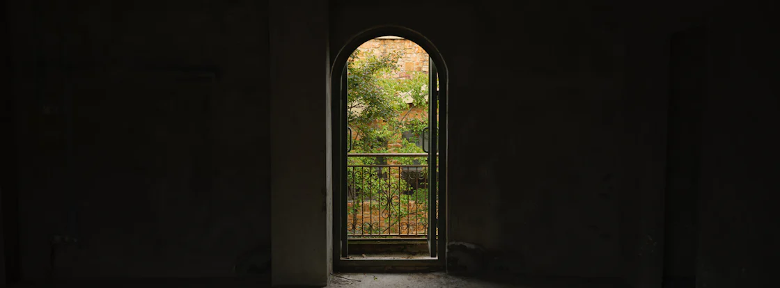 An arched doorway opening to a sunlit balcony with greenery, light spilling through shadow