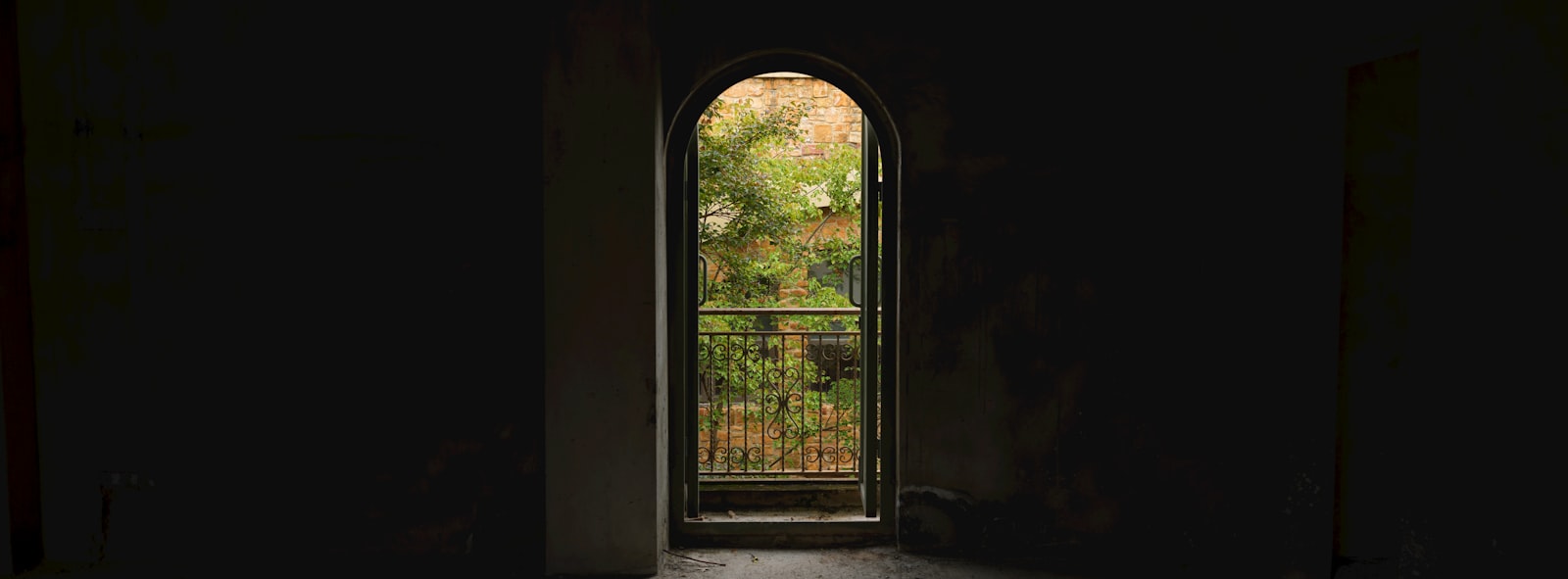 An arched doorway opening to a sunlit balcony with greenery, light spilling through shadow
