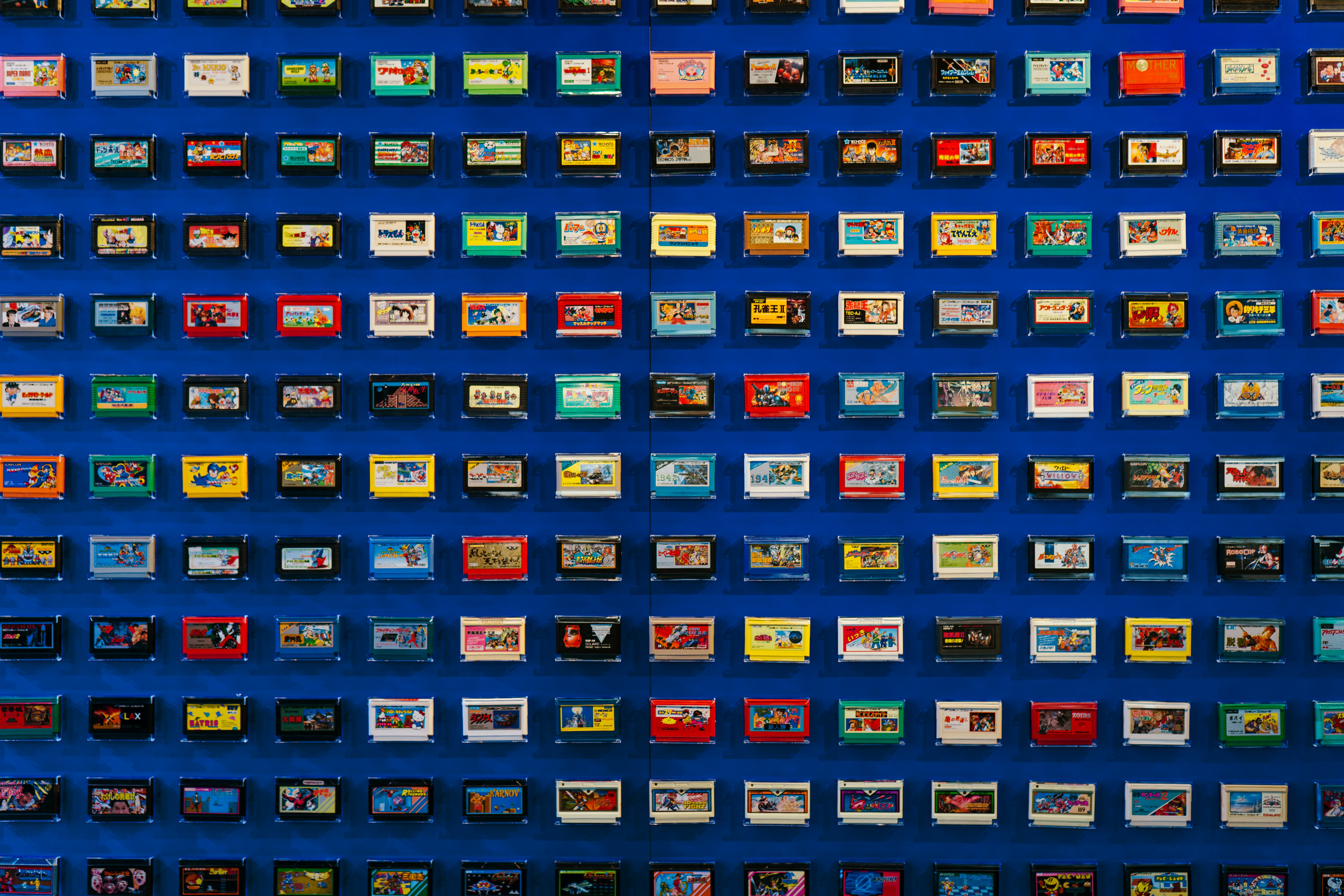 A mesmerizing grid of colorful Famicom (NES) game cartridges covers a deep blue wall in a retro gaming exhibition. The meticulously arranged collection features a rainbow of vibrant plastic casings—bright yellows, reds, blues, and blacks—each bearing unique label art that evokes the golden age of 8-bit gaming.