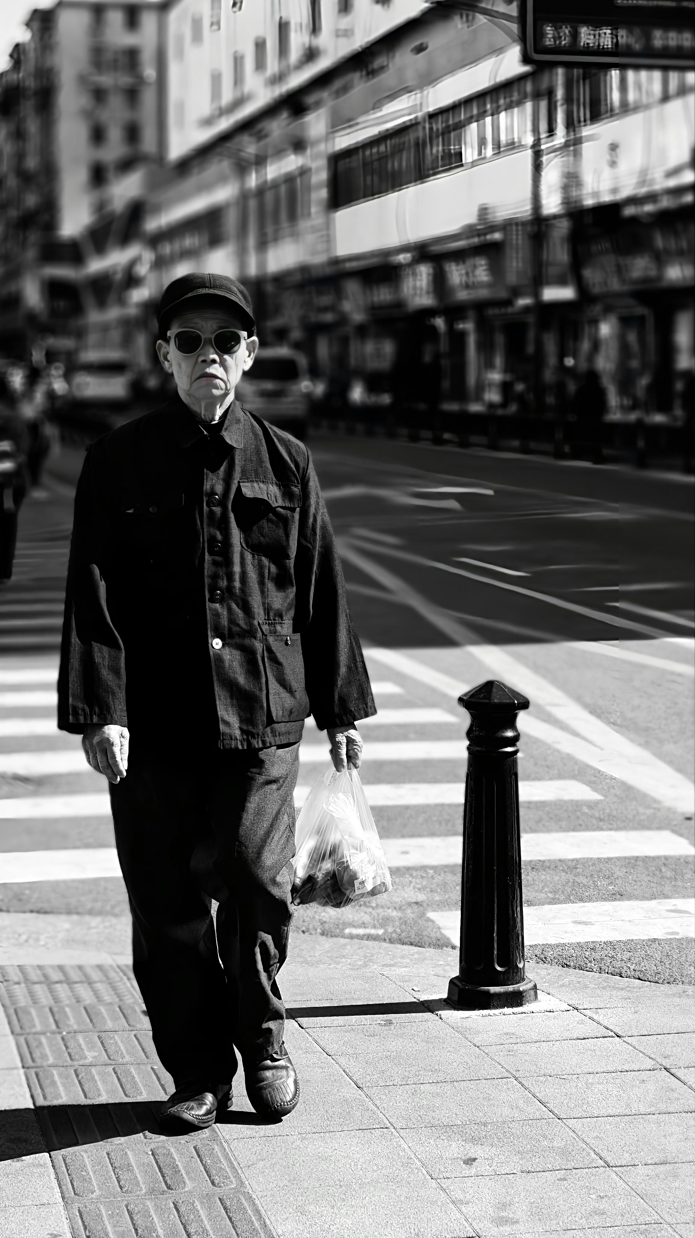 Elderly man in sunglasses walks across street carrying a bag.