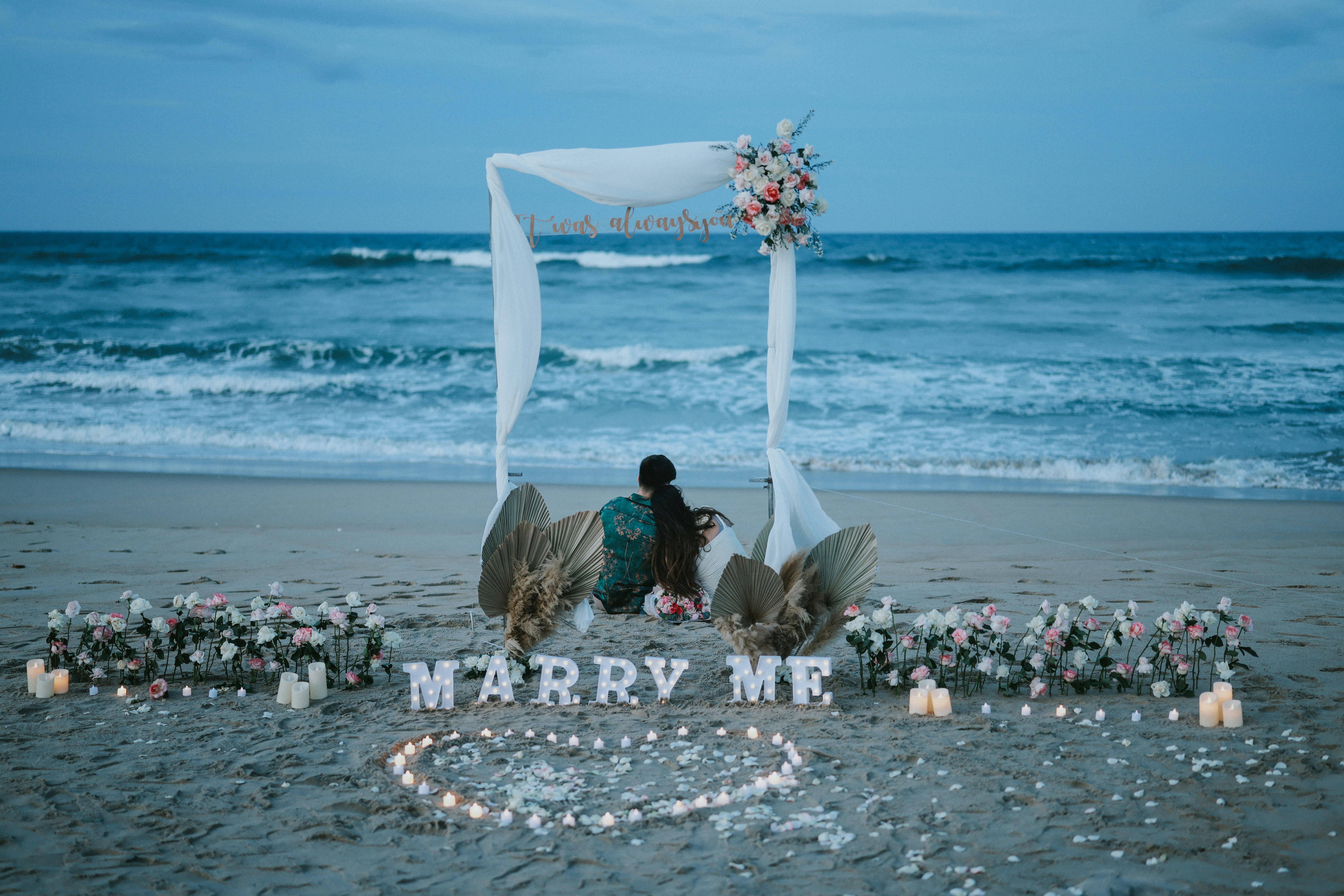 Beach proposal setup with "marry me" lights and candles.