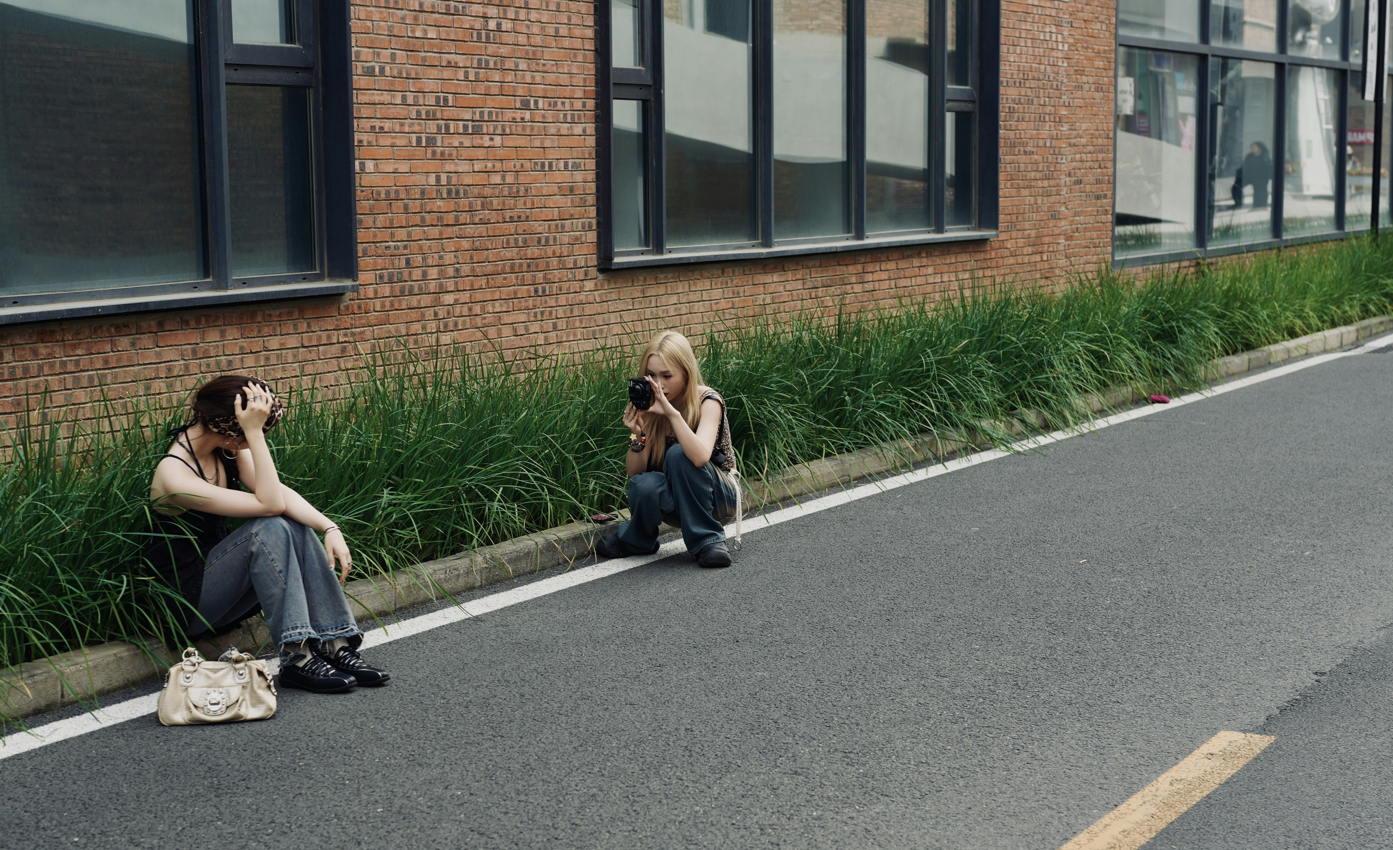 Woman photographing another woman sitting by brick building.