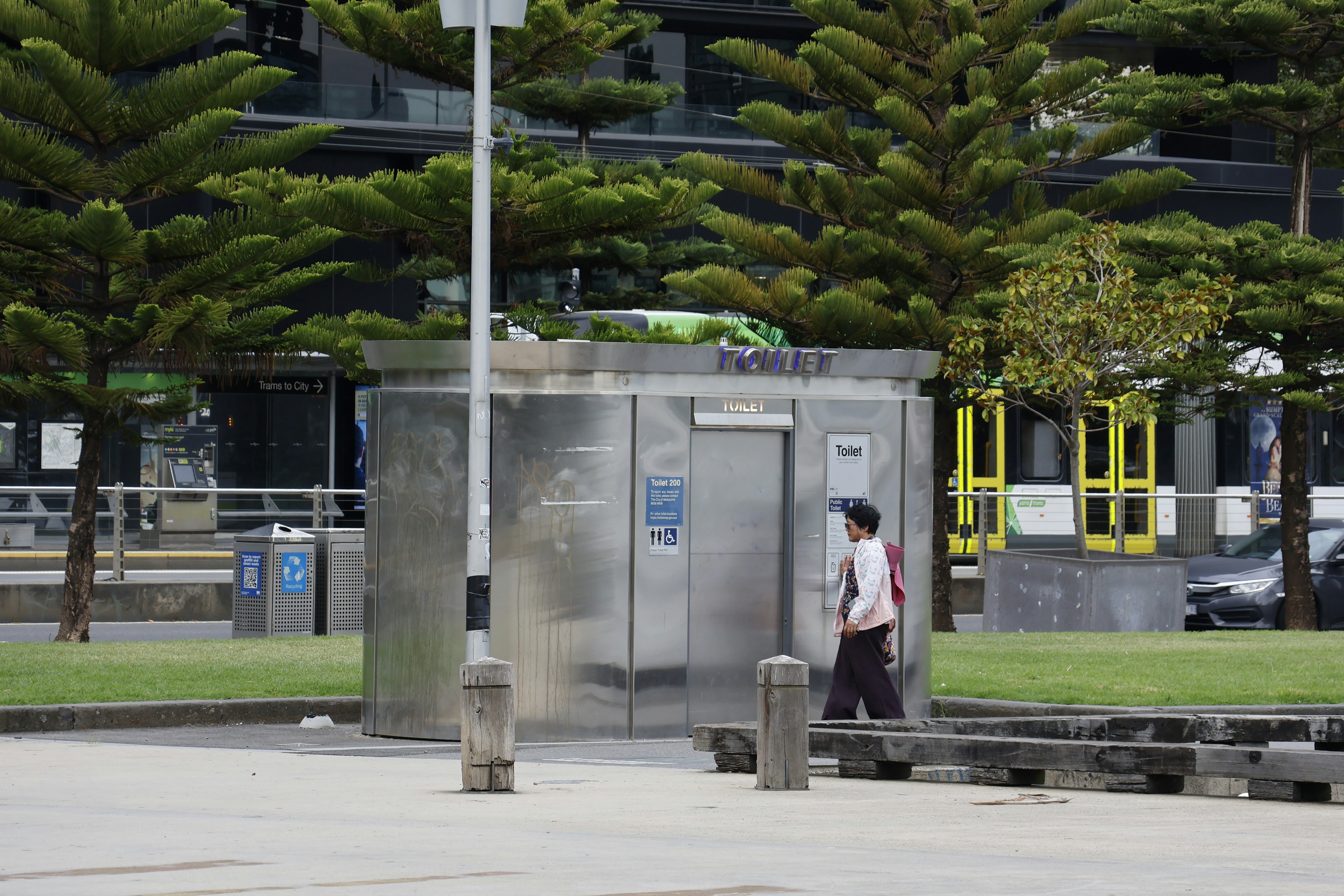 An outdoor scene featuring a modern silver public toilet, a person walking past, lush green trees, and urban infrastructure like a tram stop and bins.