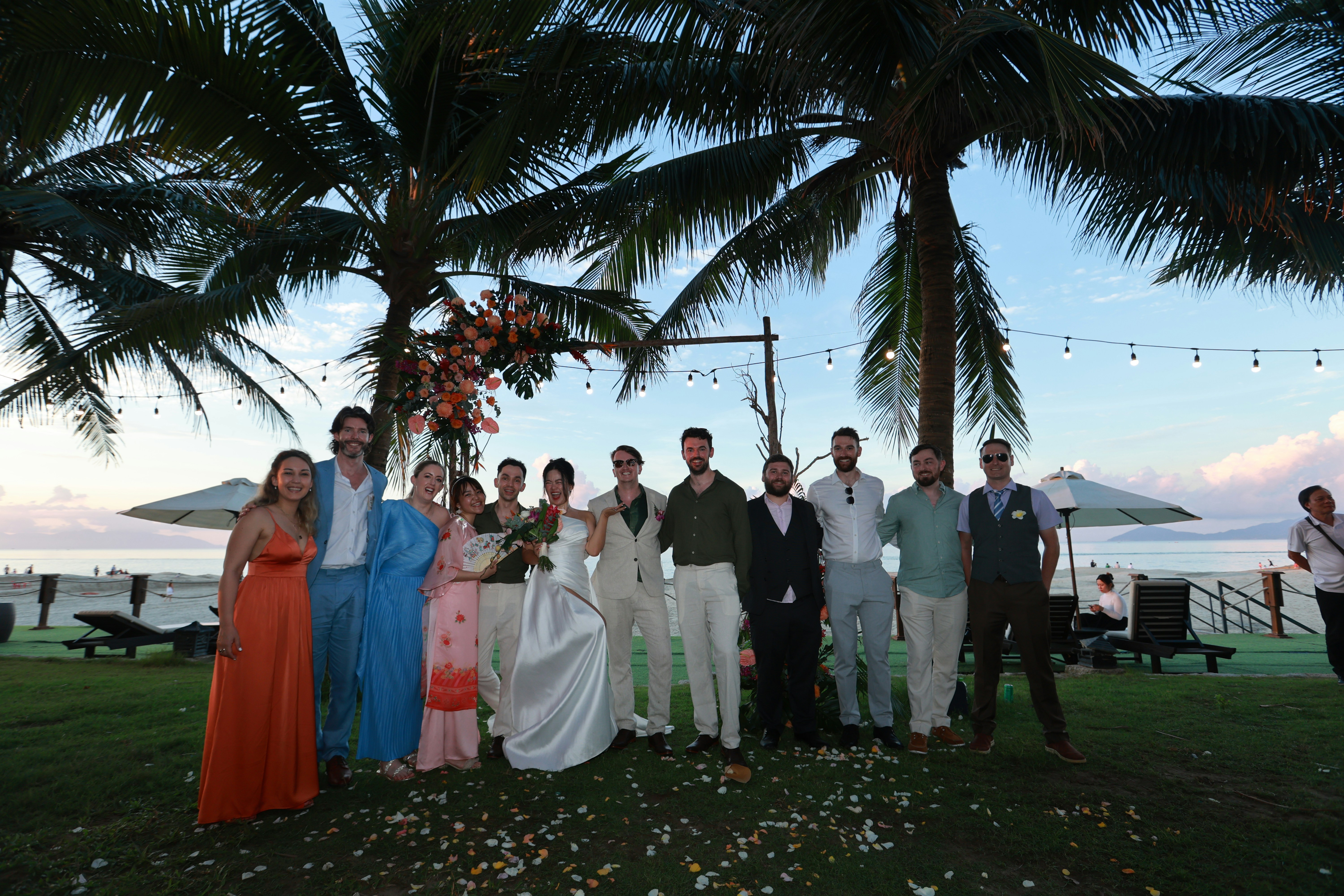 A wedding party poses under palm trees by the ocean.