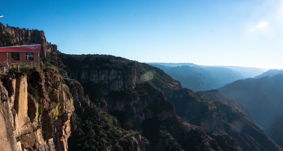 A red-roofed building on a cliff edge above the Barrancas del Cobre, with ridges fading into haze